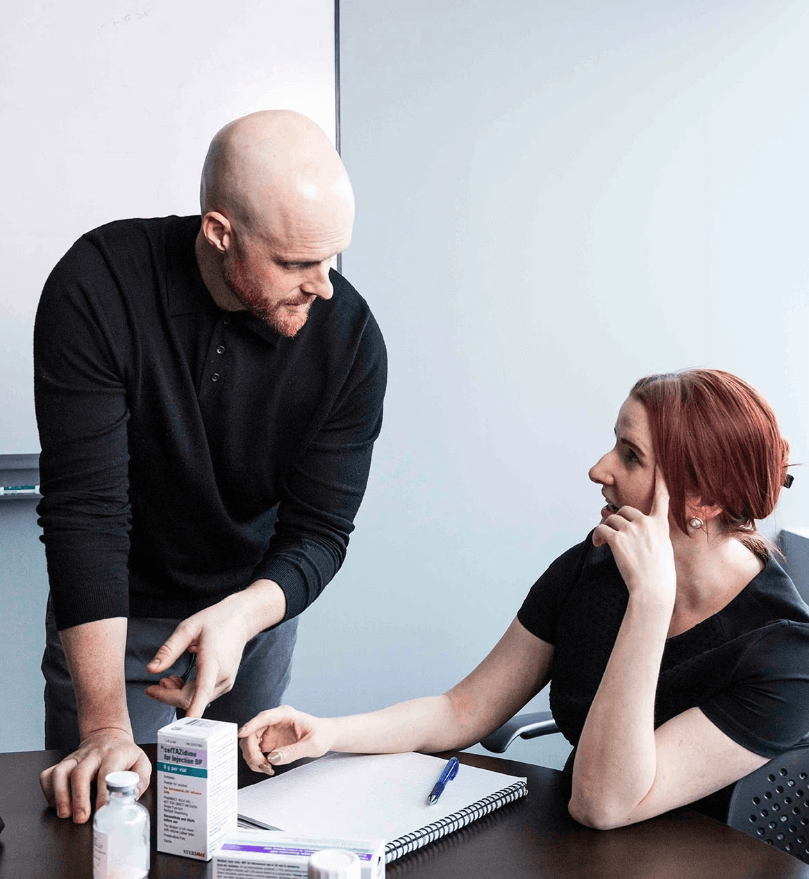 Two colleagues have a discussion at an office table with notes and pill bottles.