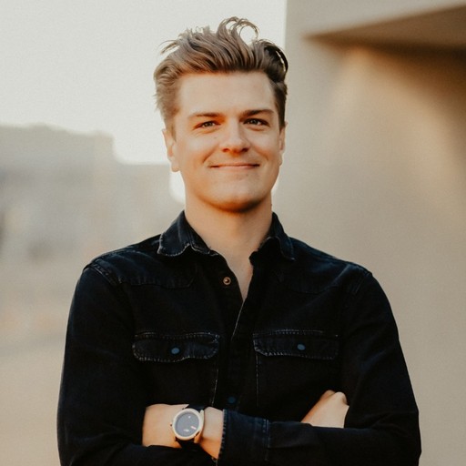 A person with light-colored hair, wearing a black button-up shirt and a watch, stands outdoors with arms crossed, smiling in front of a blurred urban background bathed in warm sunlight.