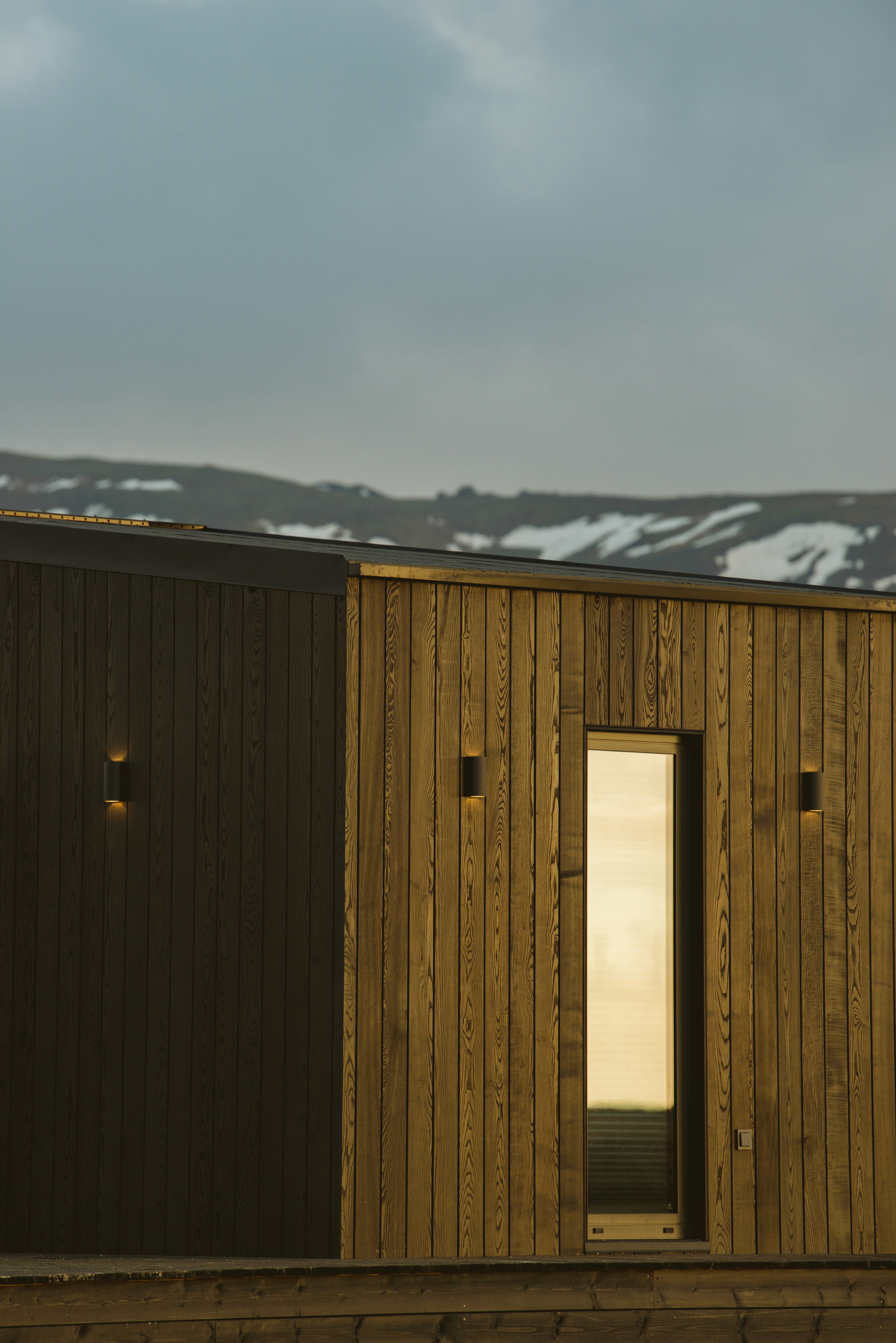 Exterior of a contemporary timber-clad cabin with vertical wooden boards and a single illuminated doorway, set against distant snow-dusted mountains beneath a calm, overcast sky.