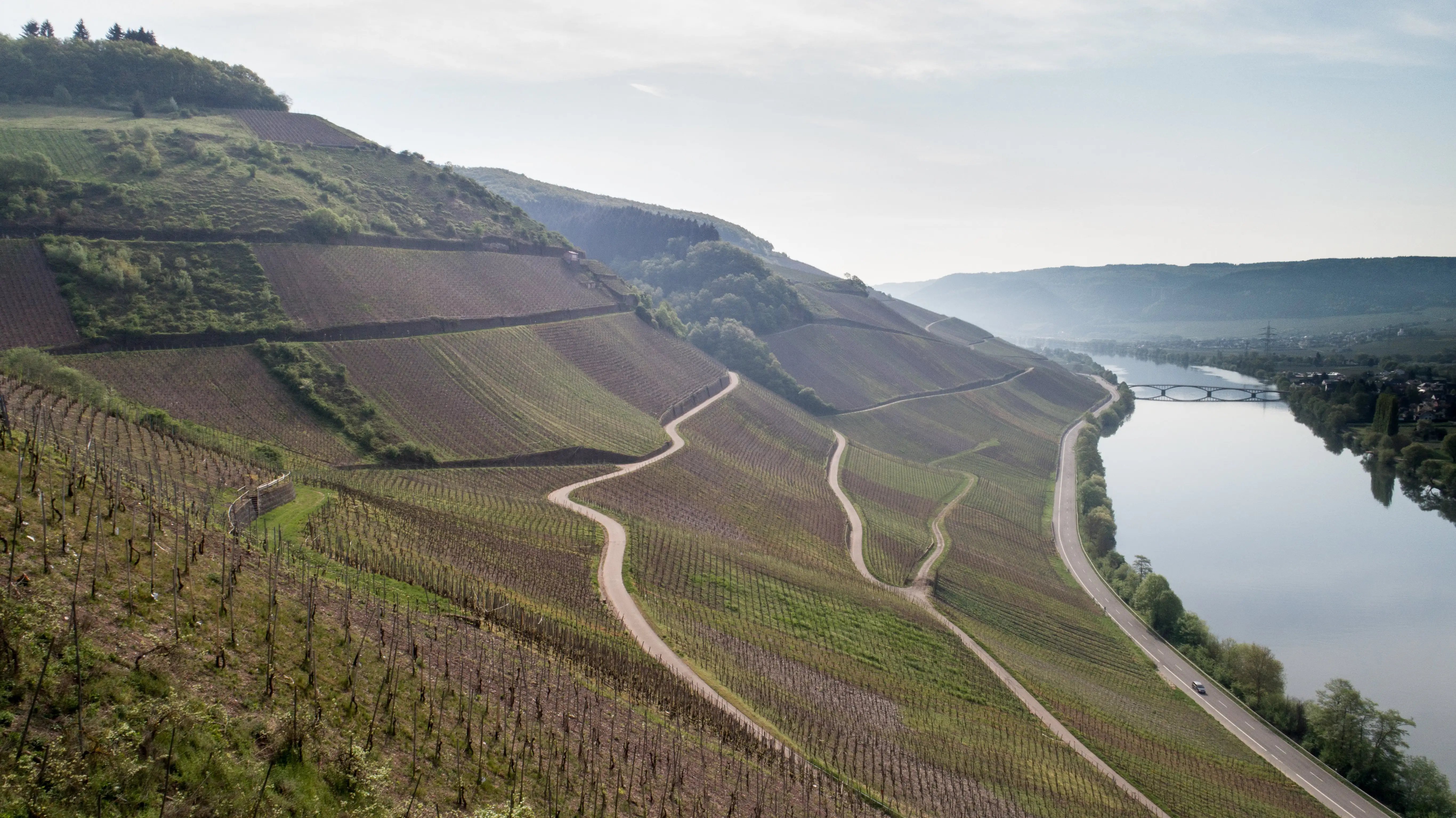 A winding road snakes up a hillside above a river, surrounded by lush greenery and a cloudy sky.