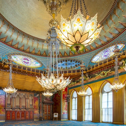 Ornate room with large, intricate chandeliers, arched windows, vibrant wall art, and a richly decorated, domed ceiling.