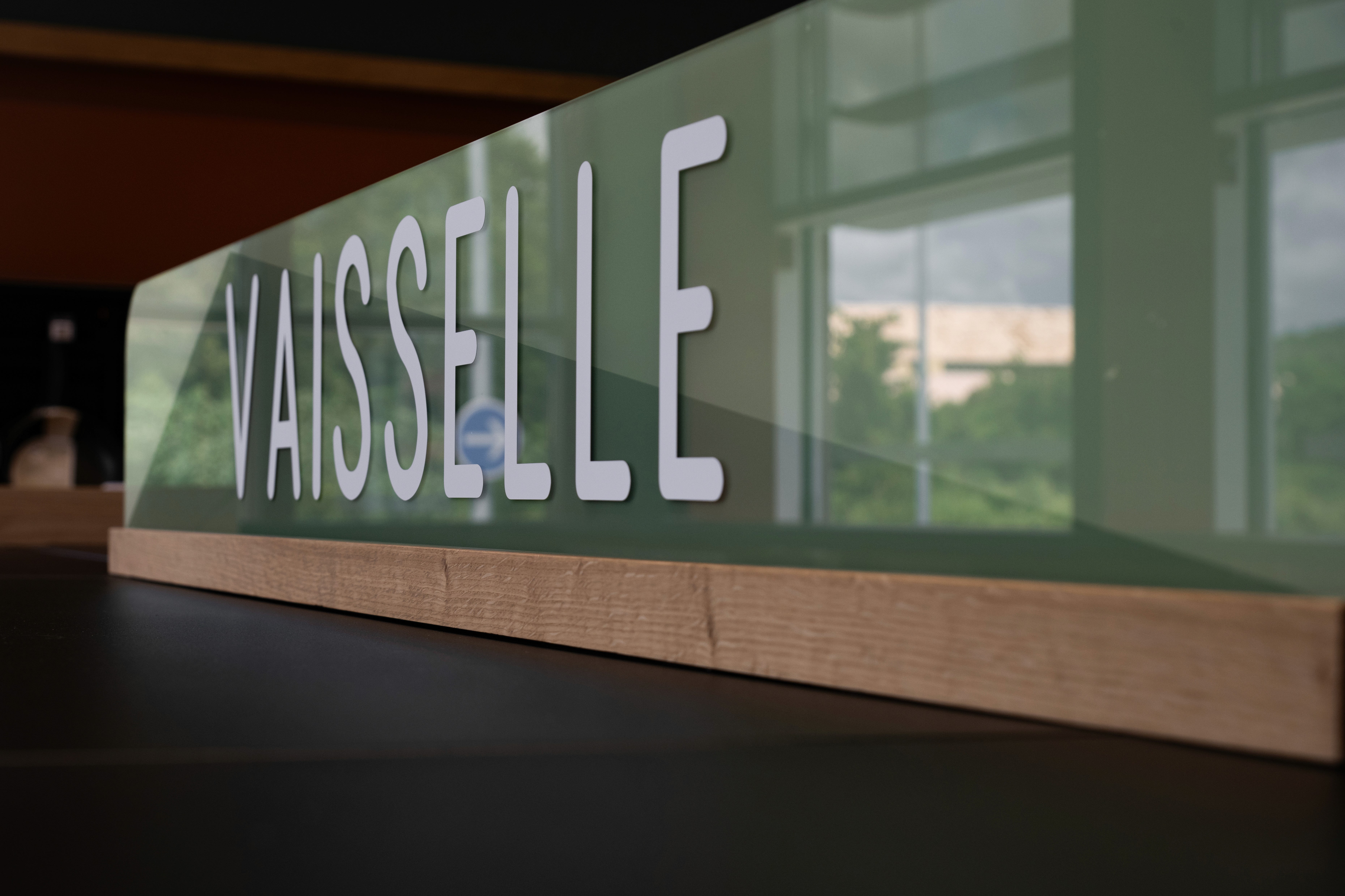 Gros plan sur une enseigne verte portant l'inscription « VAISSELLE » concu par LeGraveur signalétique en Martinique; en lettres blanches. L'enseigne repose sur un socle en bois, avec de grandes fenêtres et de la verdure visibles en arrière-plan.