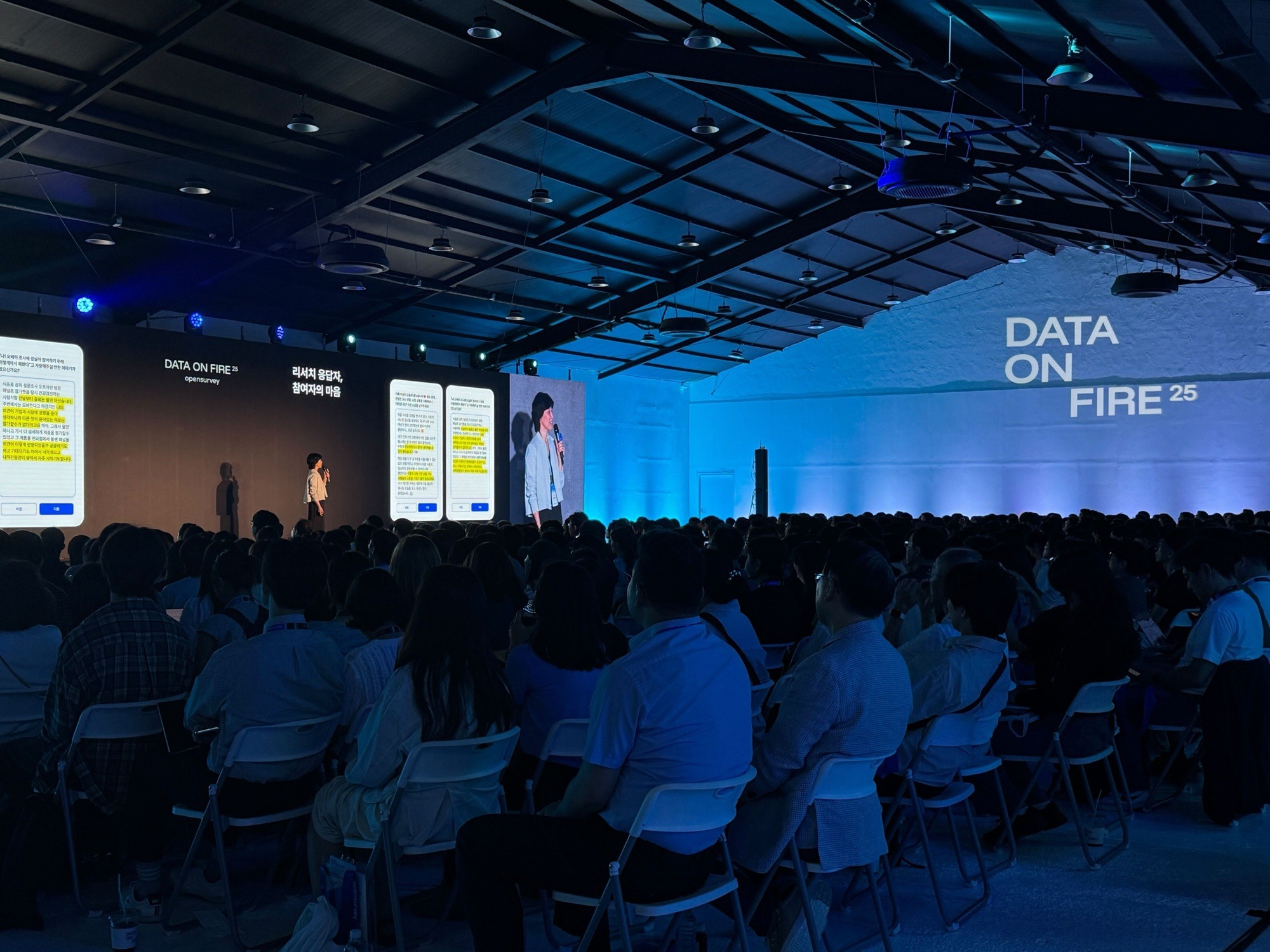 A conference scene showing a large audience under a structure, with screens displaying presentations in a blue-lit room.