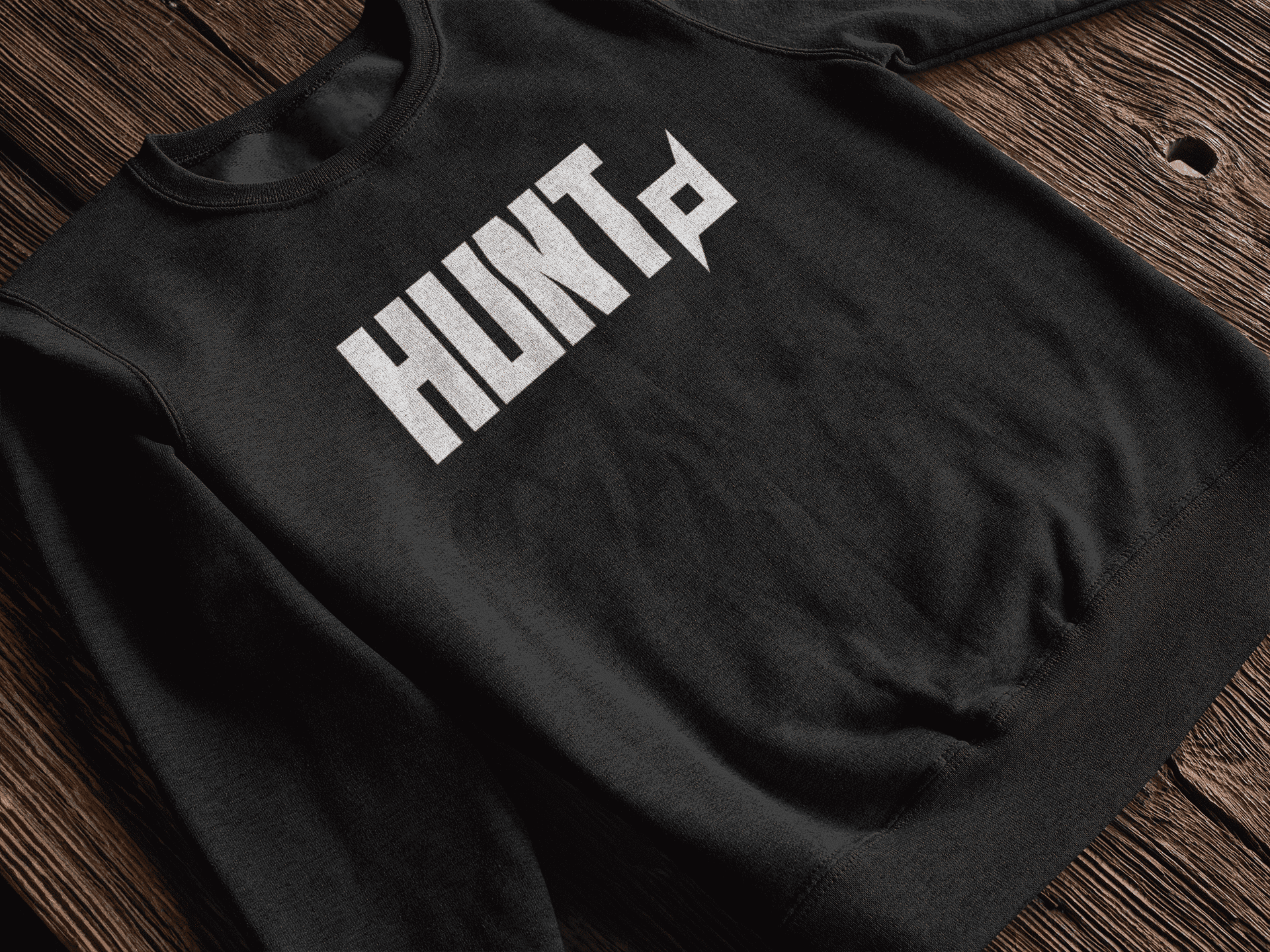 A black crew-neck sweatshirt laid out on a wooden table. The front of the sweatshirt displays the bold 'HUNT' wordmark in white, featuring a Shuriken symbol extending from the letter 'T' to emphasize motion and intensity. The textured wood surface contrasts with the dark fabric, adding warmth and depth to the presentation of the apparel.