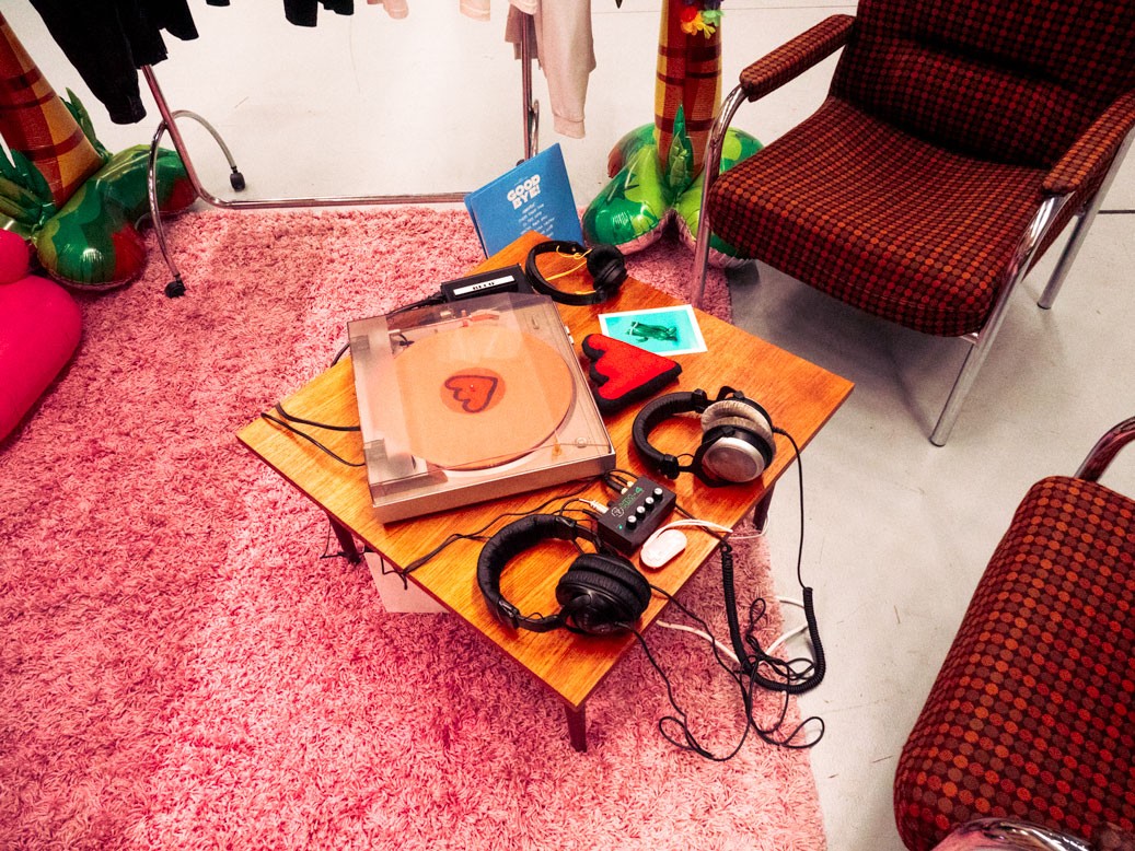 A cozy setup at a convention booth featuring a small wooden table on a pink shag rug. On the table is a vinyl record player with a heart symbol on the turntable, a pair of large headphones, an audio mixer, and additional headphones. Surrounding the table are two retro-style armchairs with red and brown checkered upholstery. In the background, inflatable palm trees and racks of clothing add to the playful and vintage vibe of the scene.