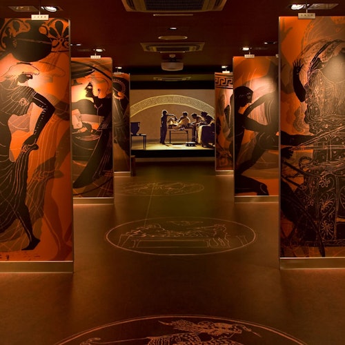 博物馆走廊两侧装饰着大型古希腊艺术展板。背景中可见一处灯光明亮的展区和参观者。