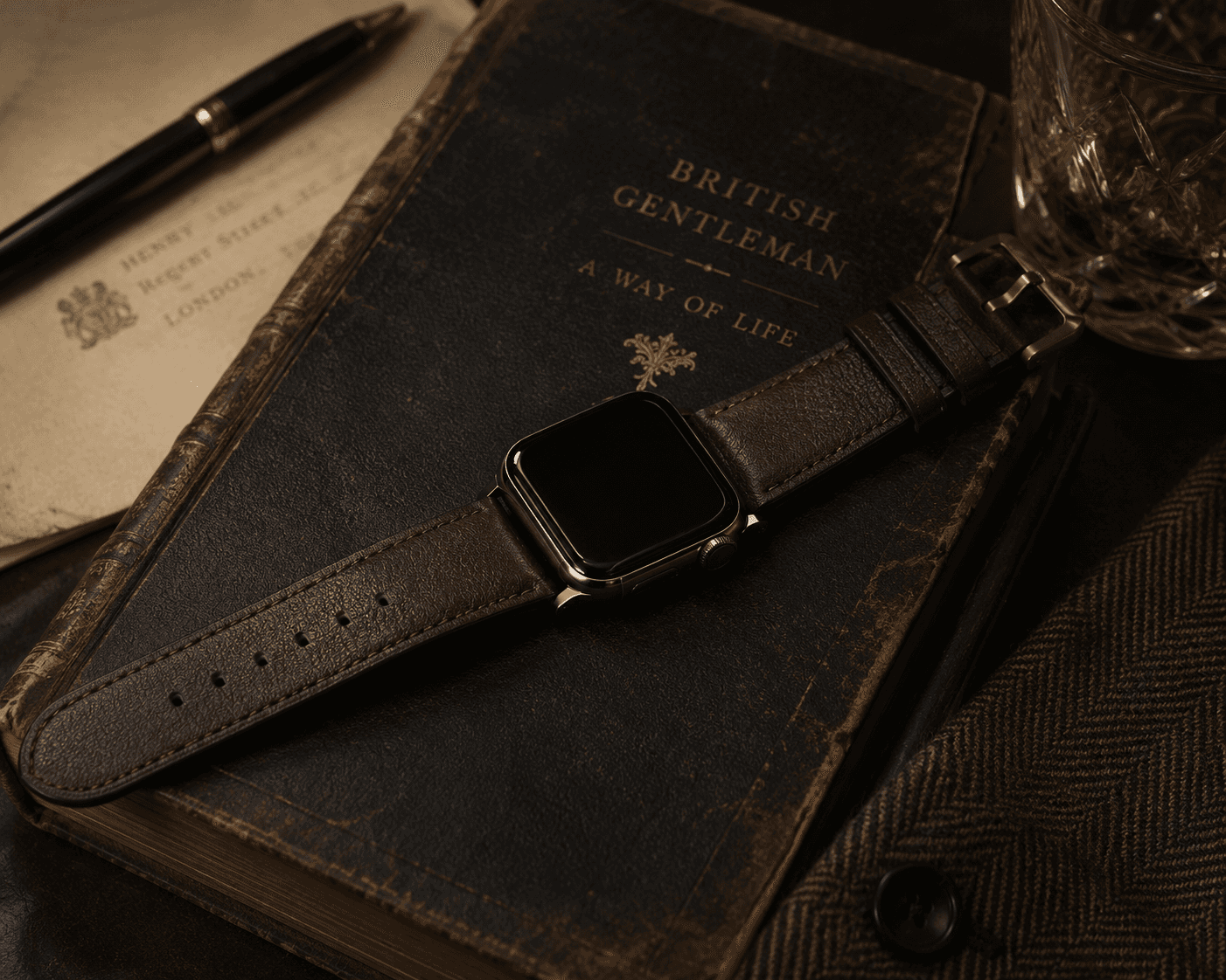 A top-down shot of a smartwatch with a dark screen fitted with a textured dark brown leather strap, lying across an antique, gold-embossed hardcover book titled British Gentleman: A Way of Life . On the left, a fountain pen sits on vintage stationery paper, while a crystal glass of whiskey and a tweed jacket sleeve frame the right edge in a warm, sophisticated desk arrangement.