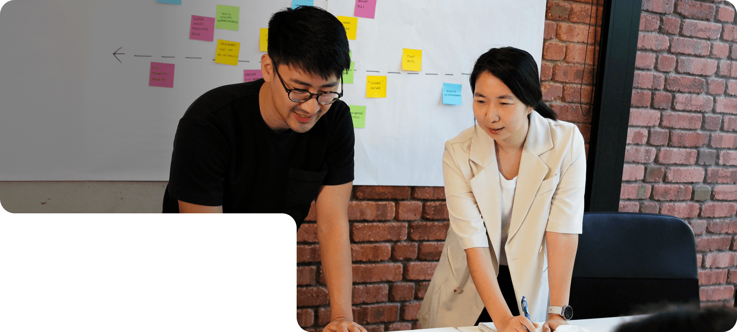 Two members of 55 Minutes collaborating in a modern workspace, discussing ideas in front of a laptop and a board with sticky notes.