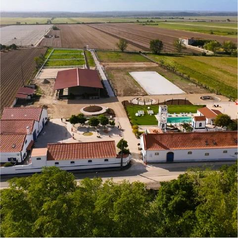 Aerial view of a Spanish countryside estate with white buildings and red roofs, green fields, a swimming pool, and a clock tower, under a clear sky.