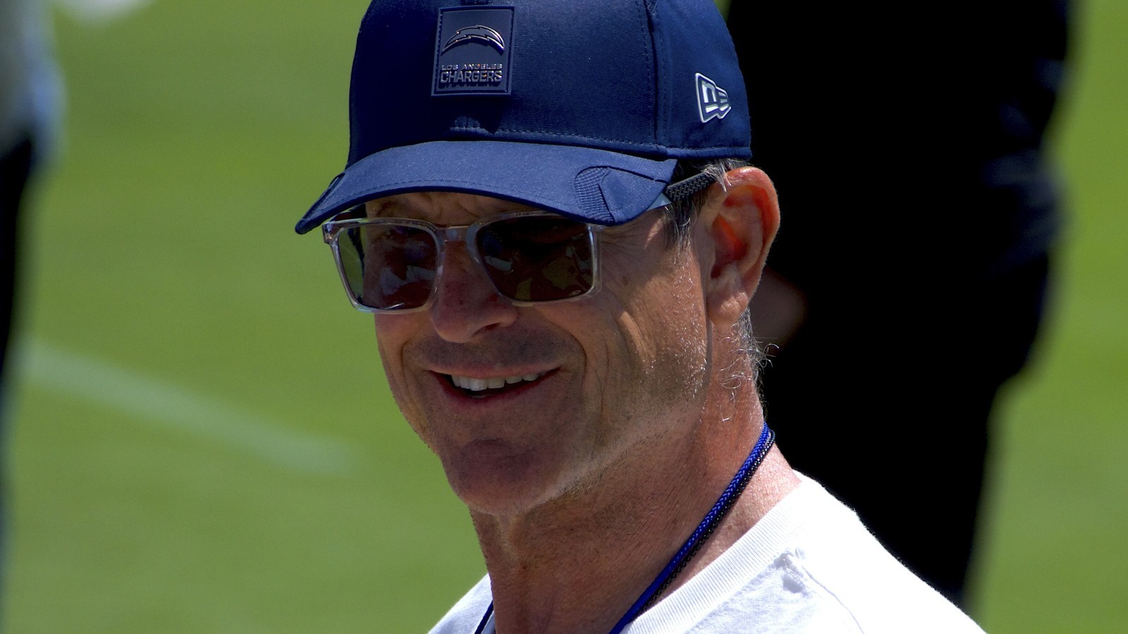 Jim Harbaugh smiling in a navy Chargers cap and sunglasses at practice