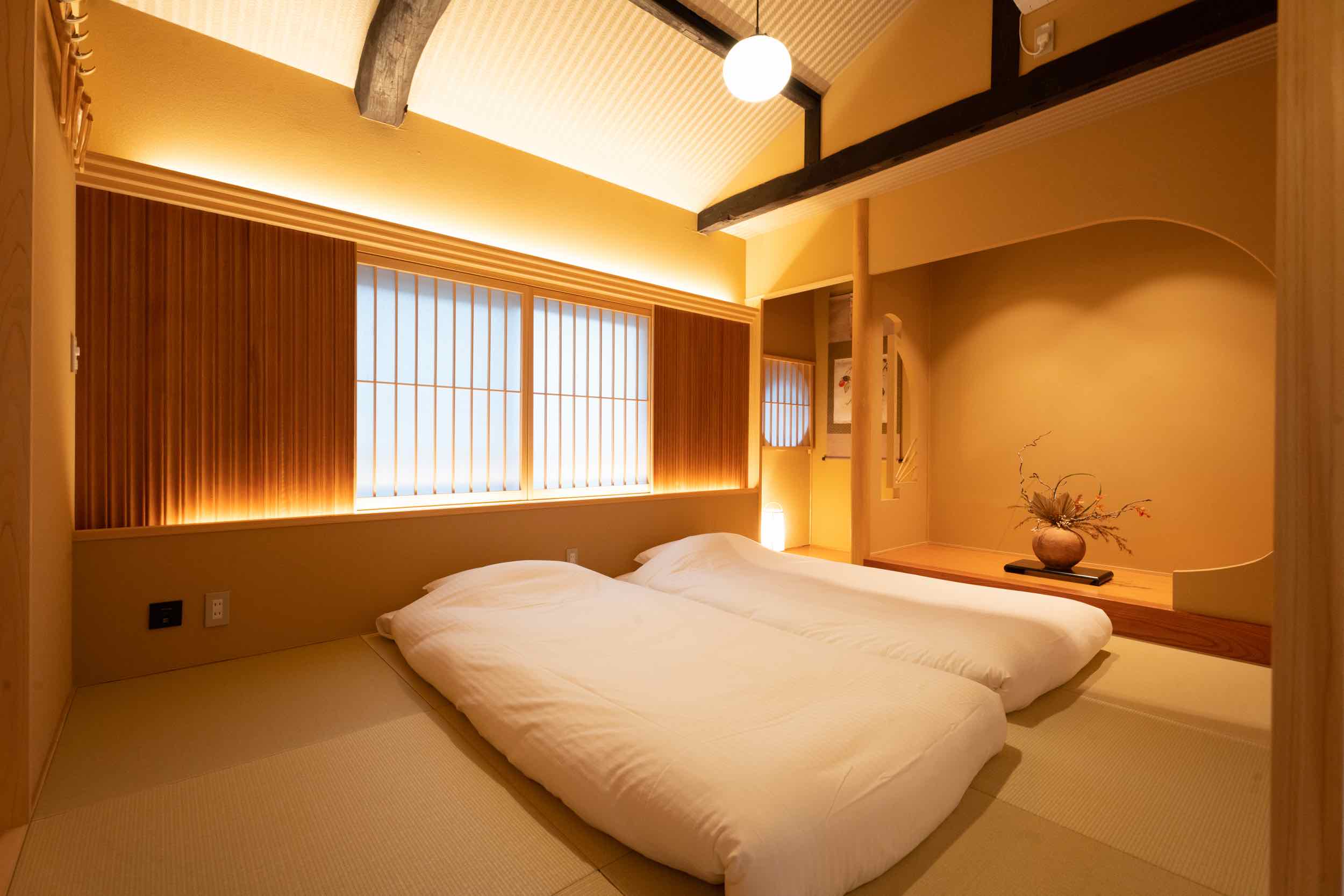 A traditional Japanese room features a minimalist design with two neatly arranged futons on a tatami mat floor, soft ambient lighting, wooden accents, and a decorative alcove with an elegant floral arrangement.