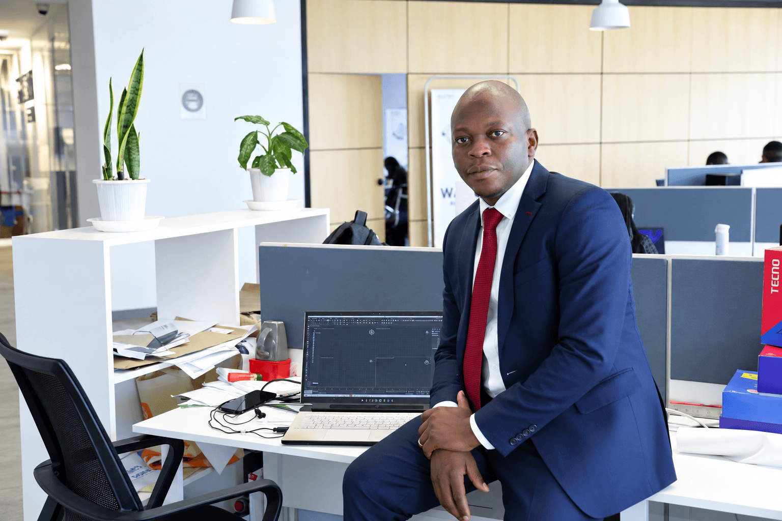 Mohammed Dabana seated beside a laptop in a modern office wearing a navy suit and red tie.
