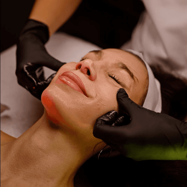 Young woman with a soft smile, resting her chin on her hands, gazing at a small serum bottle on a table