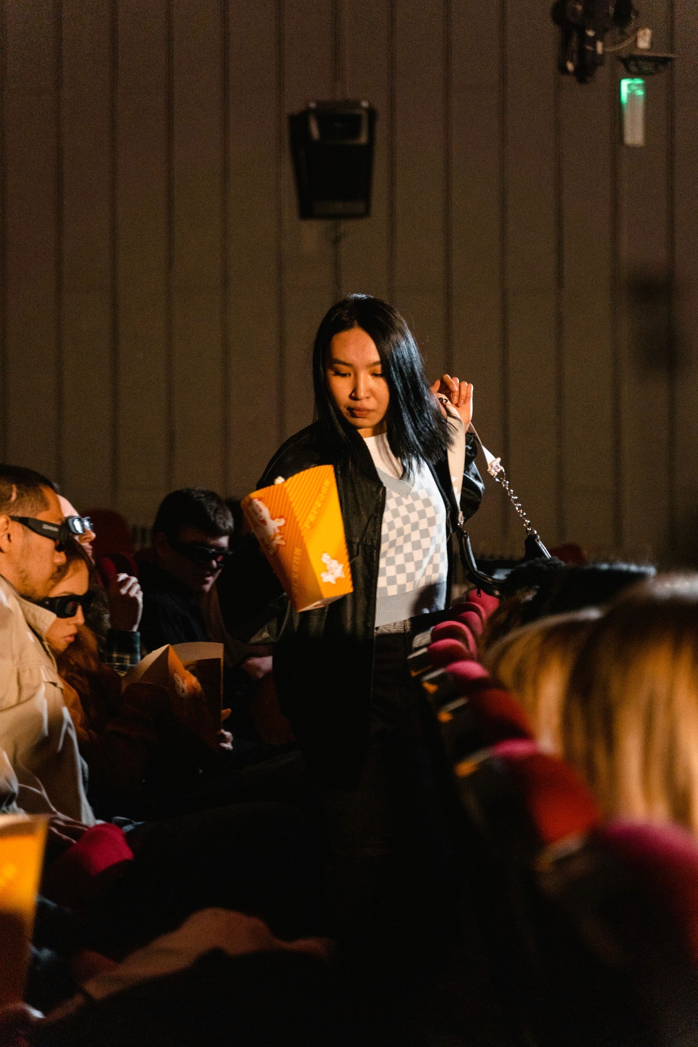 A woman carrying a tub of popcorn moves through a dim movie theater between rows of red seats before a screening.