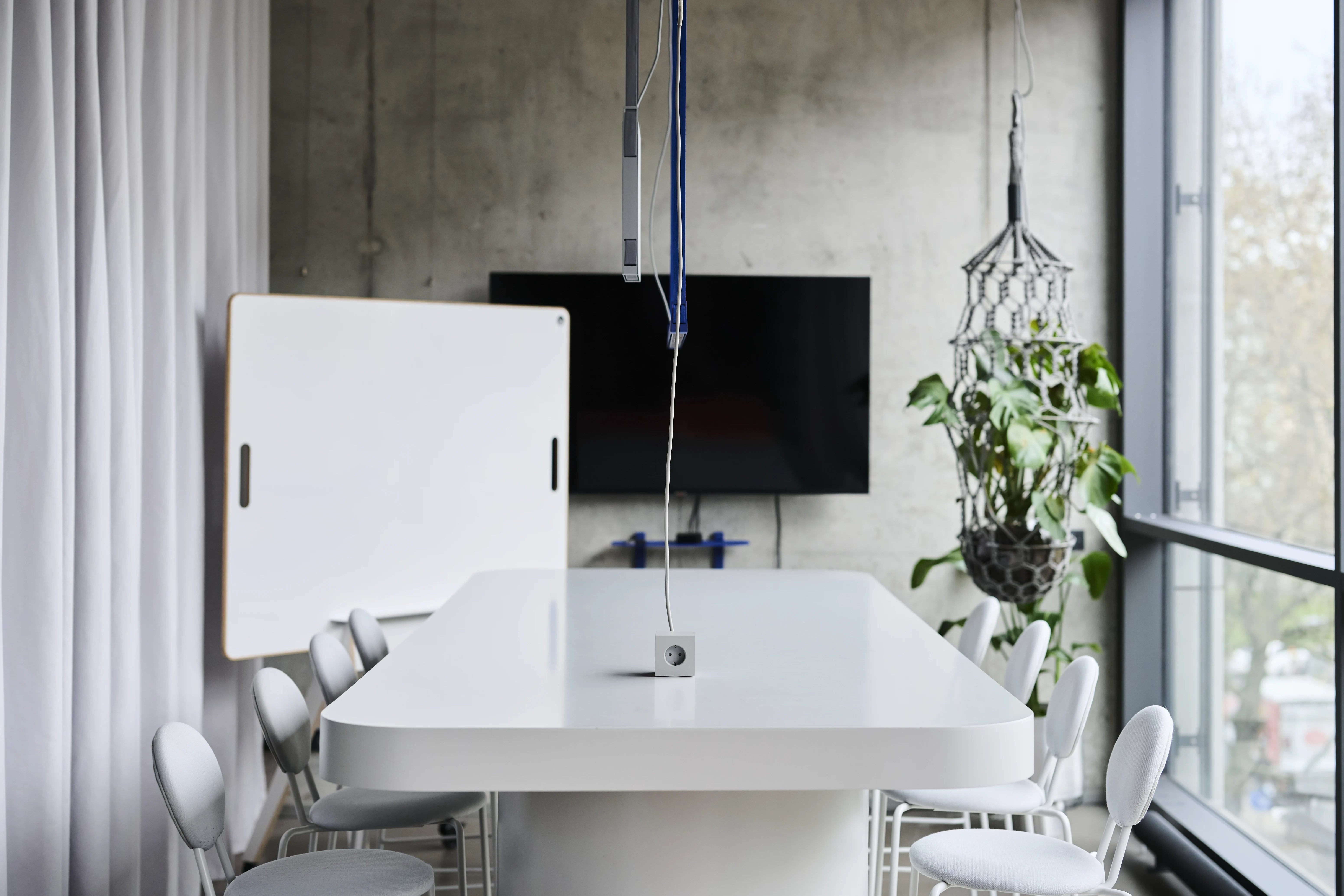 Conference table in the UNDPLUS SHOWROOM in white with light grey chairs, a mobile whiteboard, wall TV on raw concrete and a hanging plant by the window.