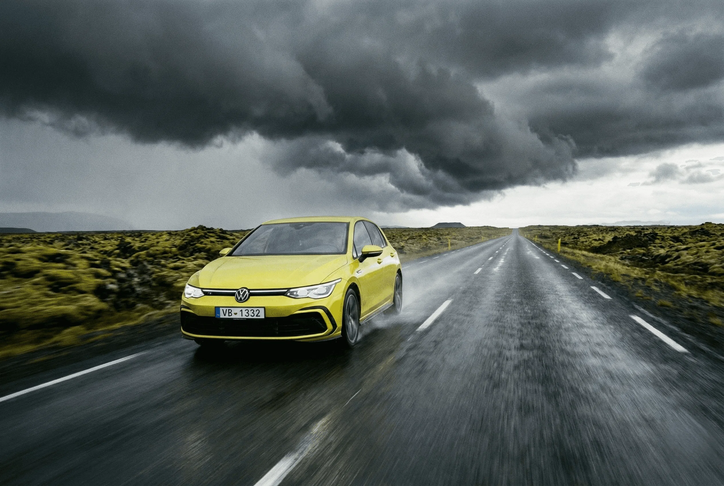 A yellow VW Golf driving on a wet road under dramatic, dark storm clouds.