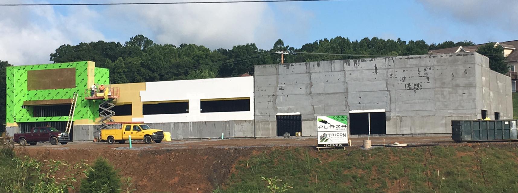 Modern commercial building under construcion with green and gray sections, surrounded by trees and clear blue sky.