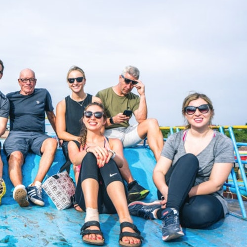 Six people sitting on a blue surface outdoors, smiling and wearing casual clothing and sunglasses.