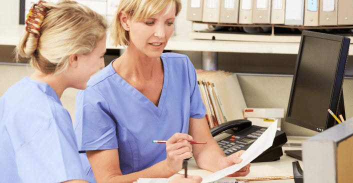 Two nurses in blue scrubs sit together at a medical workstation, reviewing paperwork, with a desktop computer, telephone, and filing binders visible in the background.
