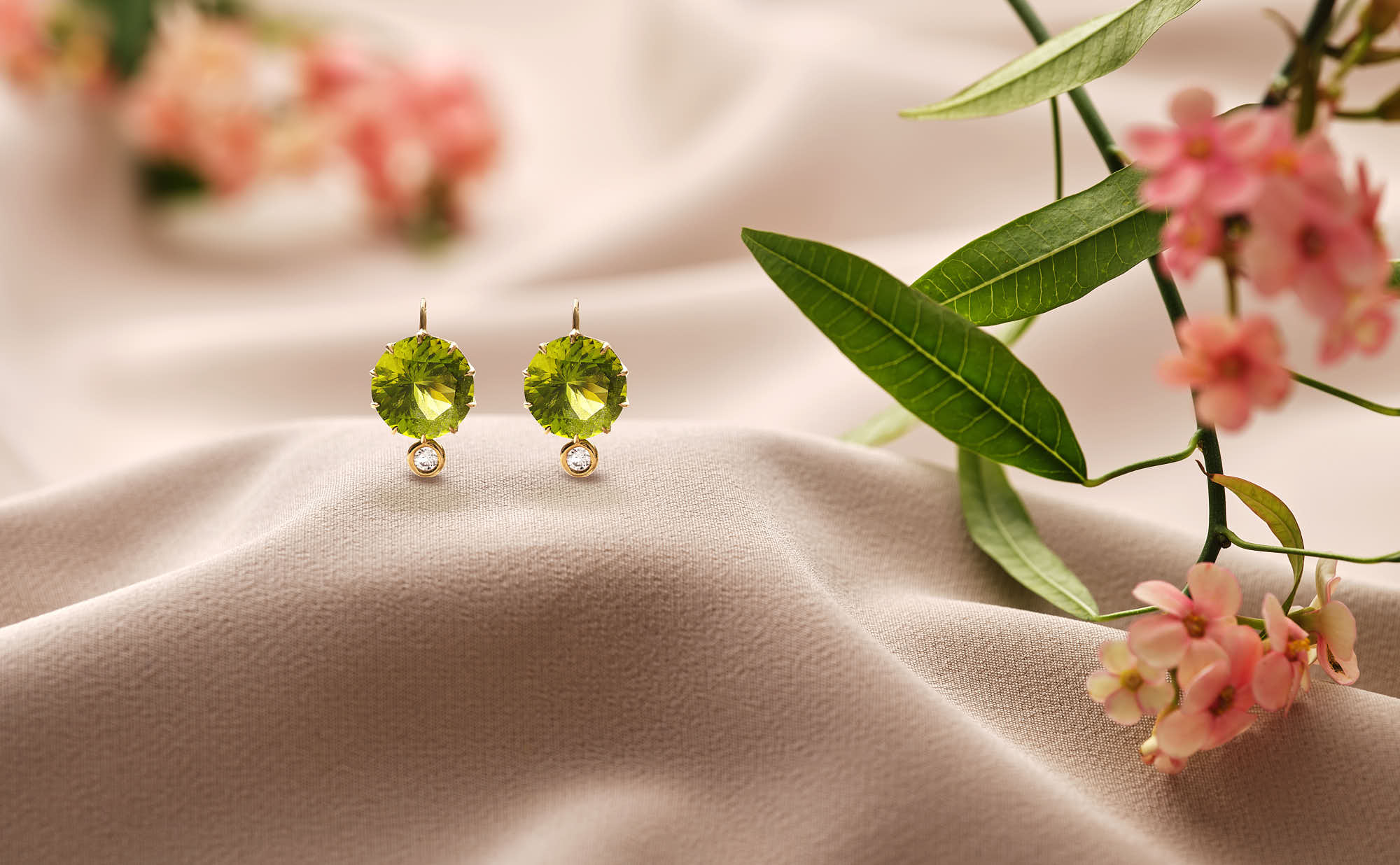 Two green earrings are placed on soft, satin fabric, accompanied by delicate pink flowers and greenery.