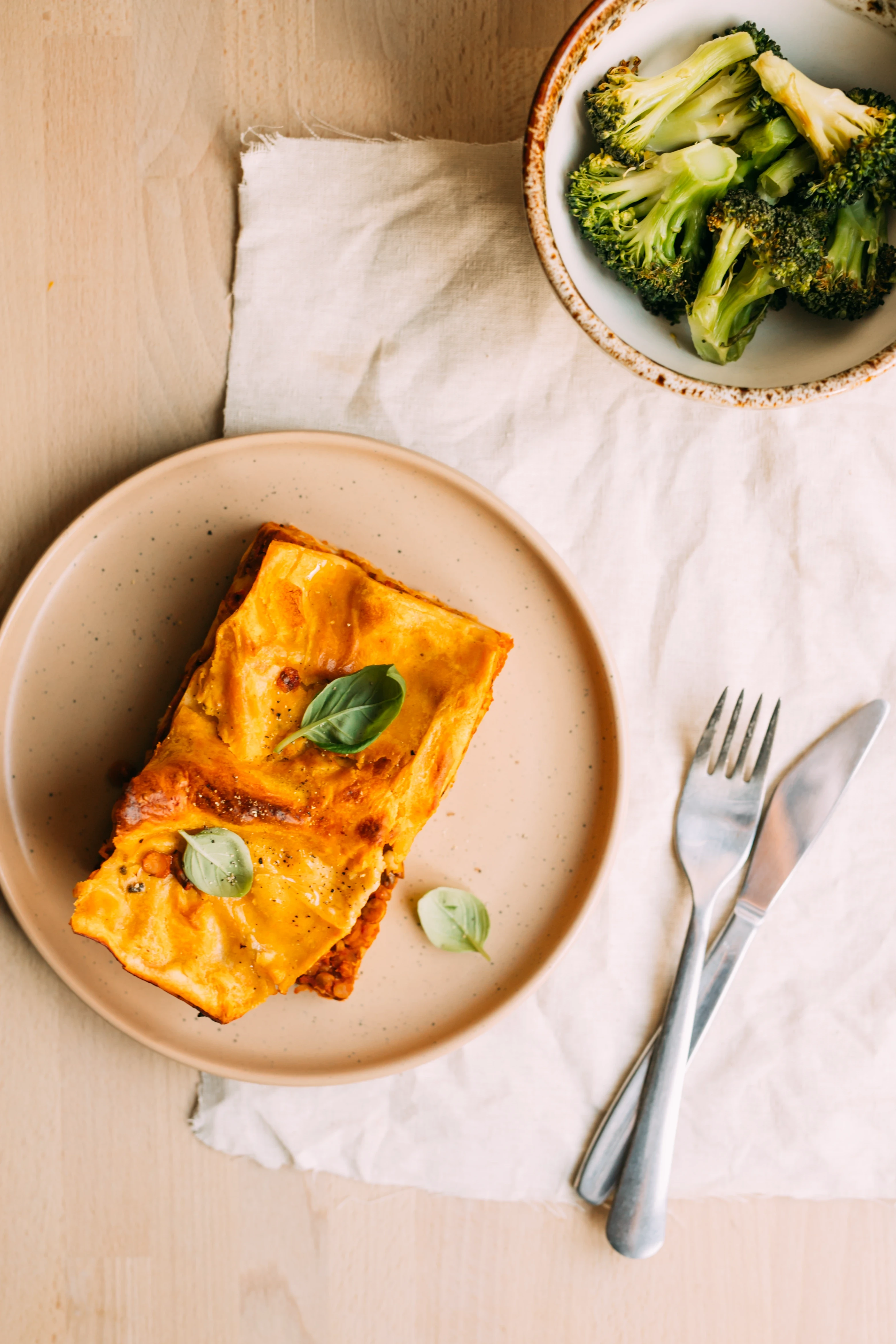 Wegańska lasagne z ragu z soczewicy podana na beżowym ceramicznym talerzu z listkami bazylii, obok miseczka gotowanych brokułów, na drewnianym blacie z lnianą serwetką