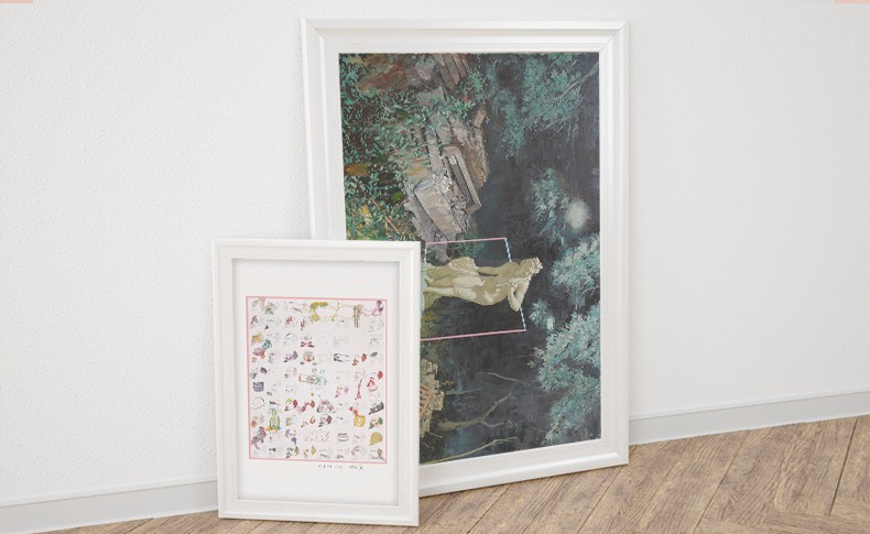 Two framed artworks lean against a white wall. The larger artwork depicts a statue in a forested setting with rocks and foliage. The smaller artwork is a grid of colorful illustrations on a white background. Both frames are white and rest on a light brown wood floor.