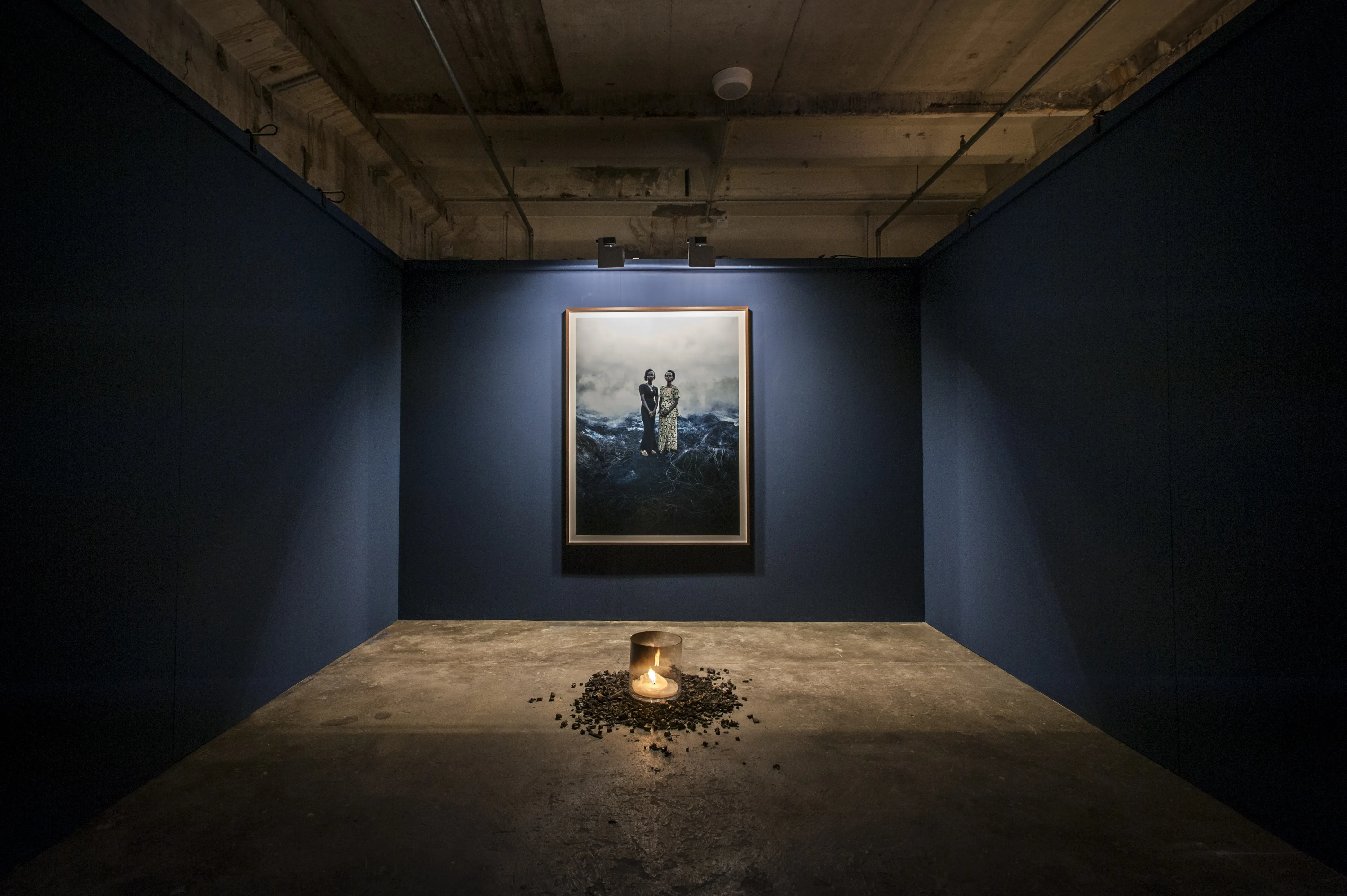 Dark blue gallery room at BERLIN PHOTOWEEK with a single spotlit photograph and a glowing vessel on charcoal on the concrete floor.