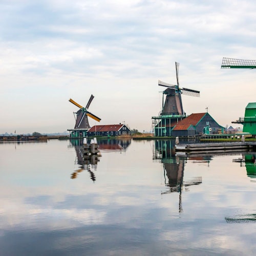 Three traditional windmills near the water, reflecting on a calm surface. Red and green buildings surround them under a cloudy sky.