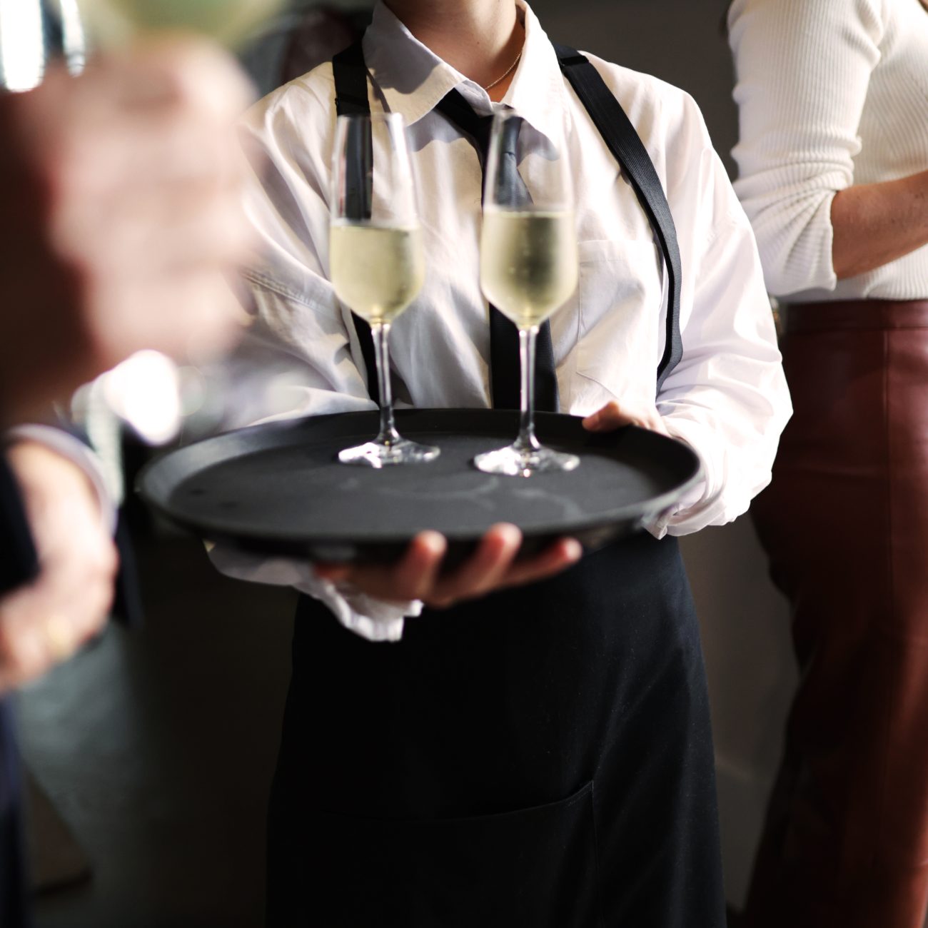 Waiter in a bow tie and apron carrying a tray of champagne glasses through a crowd