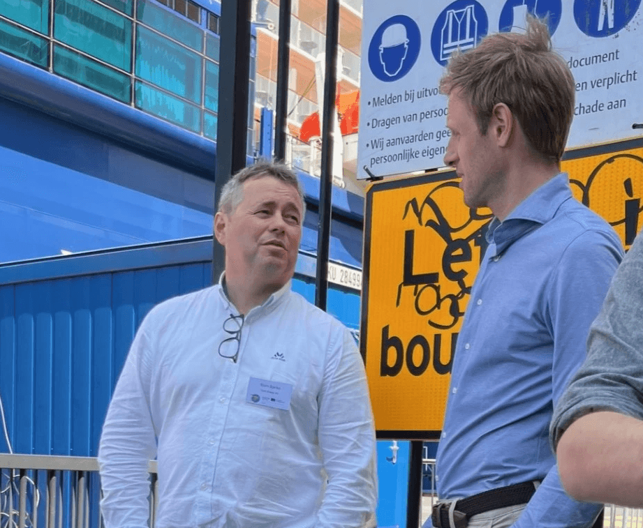 Two men in casual dress converse in front of a colorful backdrop with signs and construction elements.