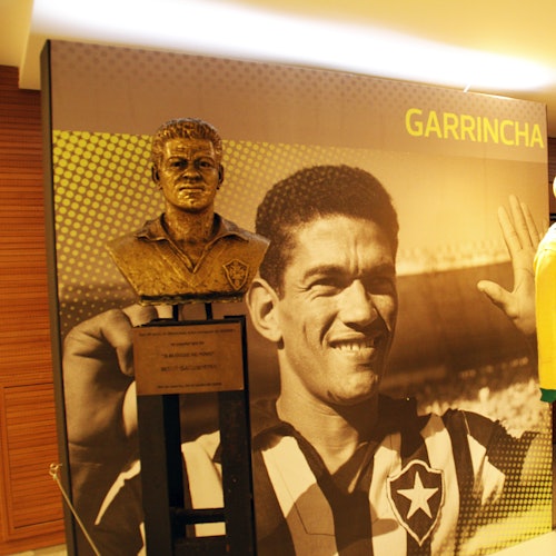 Un bust din bronz al lui Garrincha în fața unei fotografii mari în care el zâmbește purtând un tricou de fotbal.