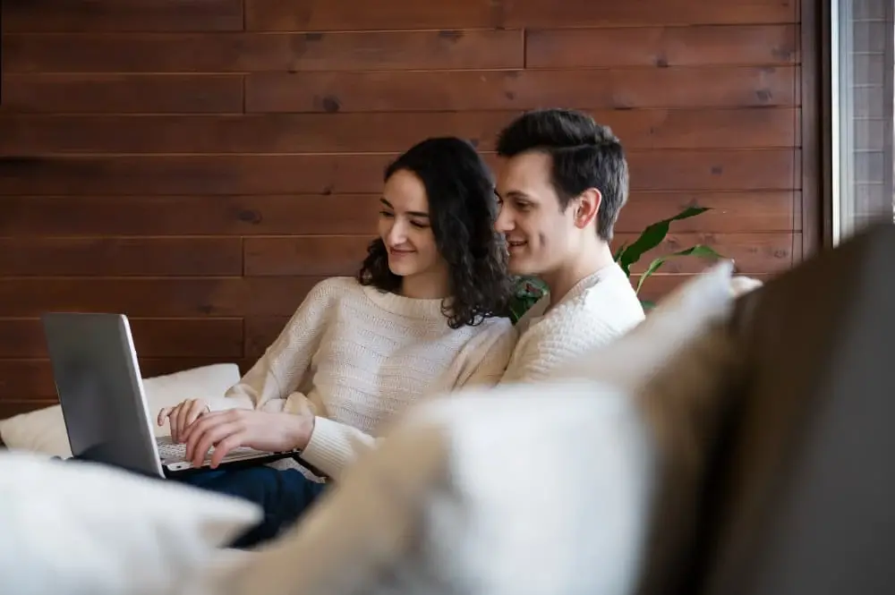 A couple sits together on a cozy sofa, smiling as they browse on a laptop in a warm, wood-paneled room. The image conveys comfort, connection, and the ease of comparing investment loan options through Chris Lewis Home Loans.
