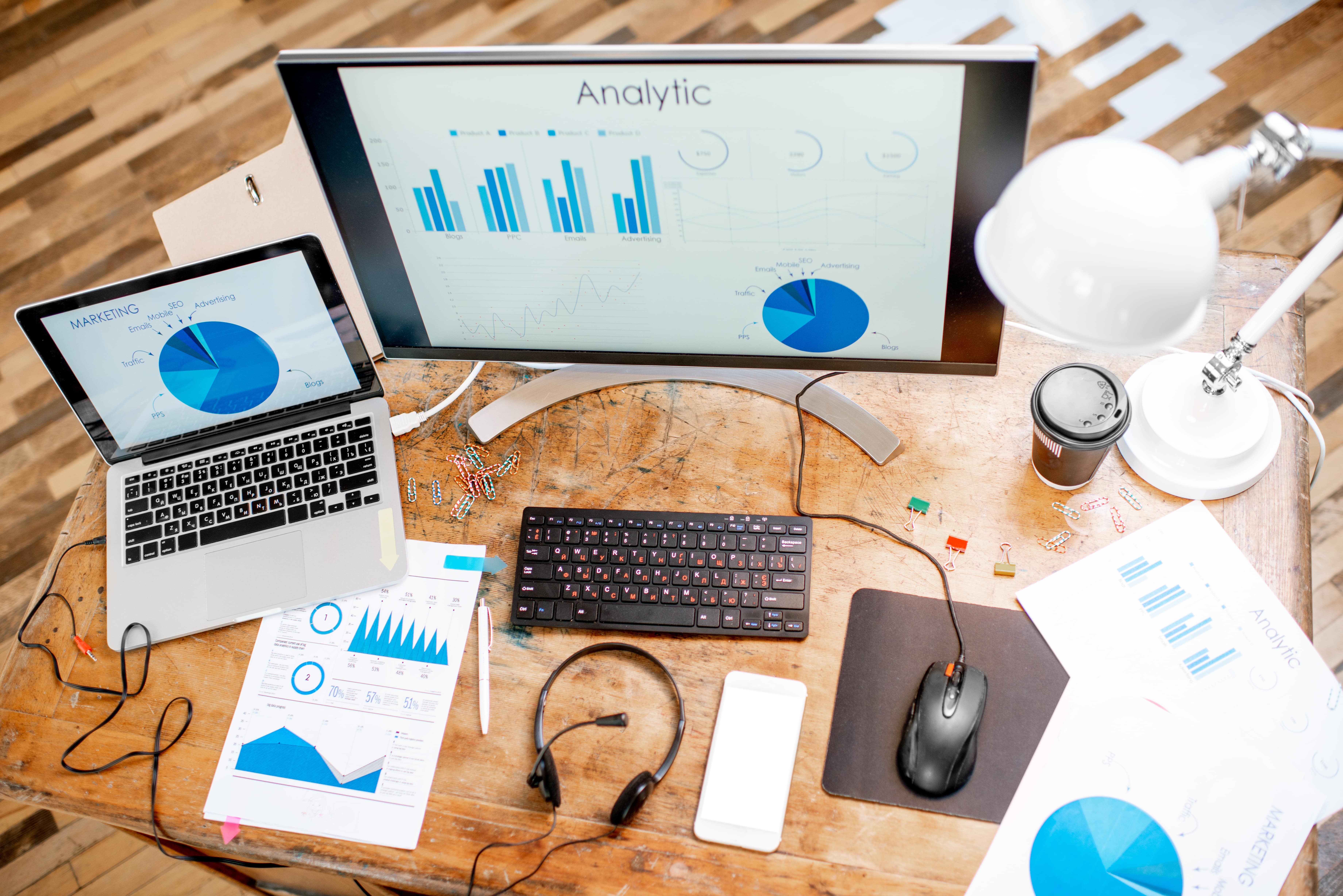 A top-down view of a data analyst's wooden desk, featuring a laptop and a large monitor displaying marketing analytics charts and business performance graphs. The workspace is equipped with a headset, keyboard, mouse, and printed reports, illustrating a data-driven work environment.