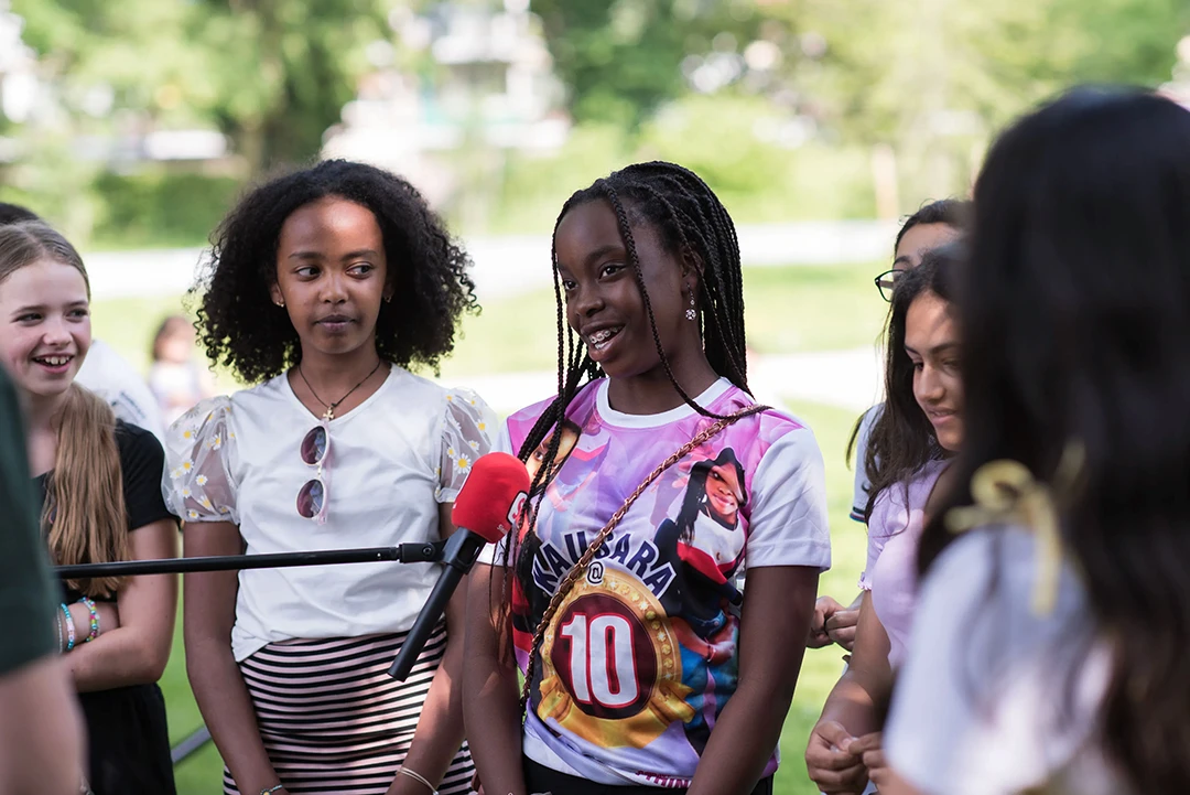 Meisje spreekt in een microfoon tijdens een buitenactiviteit, omringd door leeftijdsgenoten die glimlachen en luisteren