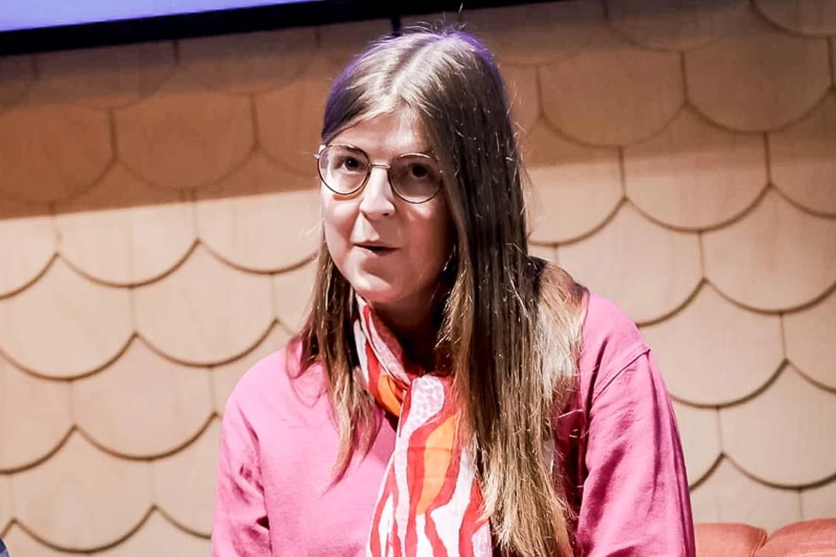 A panel speaker wearing a pink top and patterned scarf sits on stage under bright lighting. The textured wall behind them suggests a formal talk or conference setting.