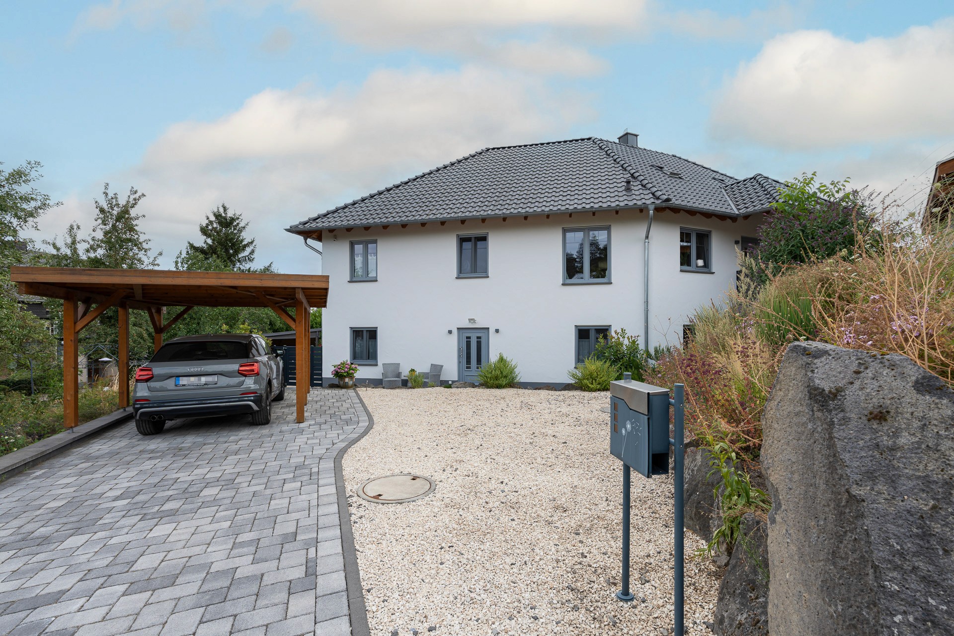 Zweigeschossiges Einfamilienhaus mit weißer Fassade, dunklem Walmdach, Holzcarport und gepflasterter Zufahrt.