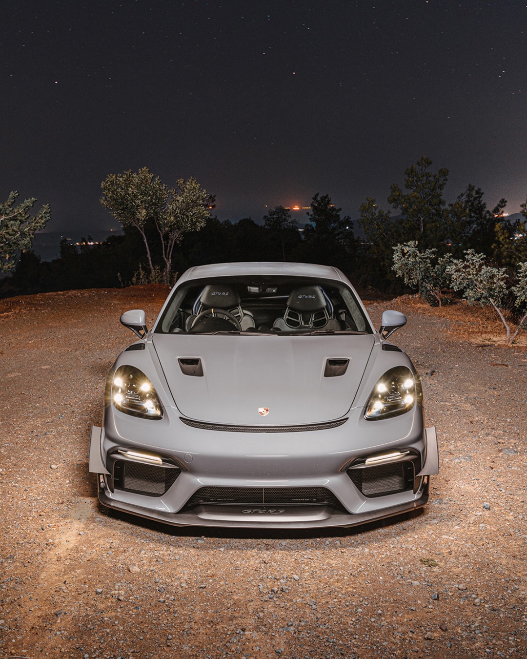 Grey Porsche 718 Cayman GT4 RS photographed head-on at night, headlights lit, showing the carbon front splitter, bonnet vents and "GT4 RS" front lip lettering, against a starry sky and distant hillside lights in Cyprus.