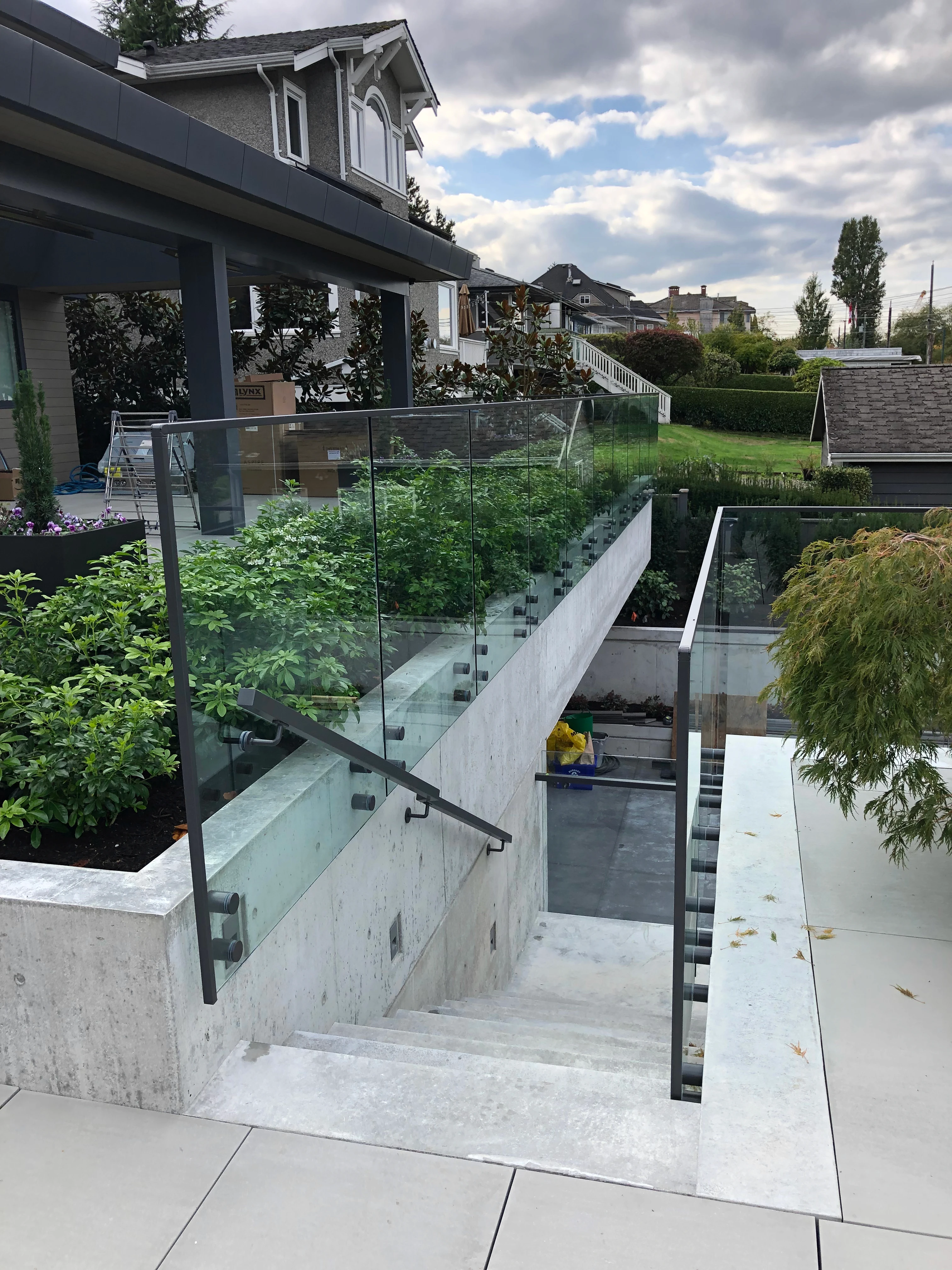 Side-mounted standoff glass railings with a dark metal handrail bordering an outdoor concrete staircase that leads to a lower level.