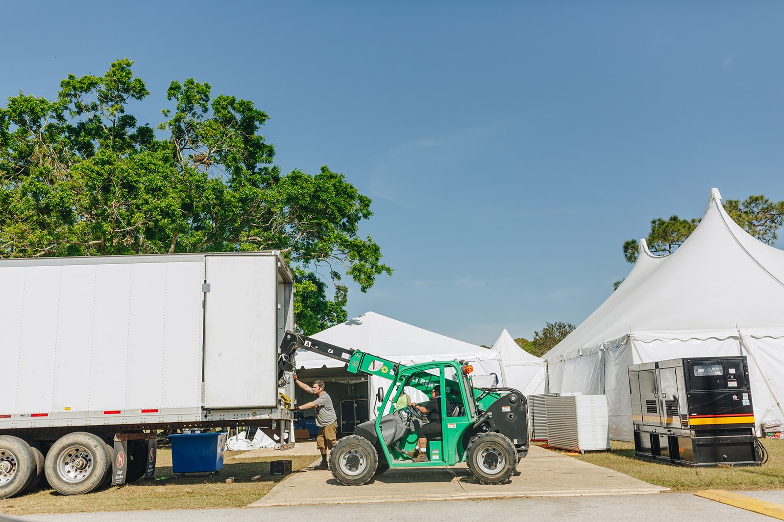 A green tractor parked beside a white truck in an open area with tents and clear blue sky in the background.