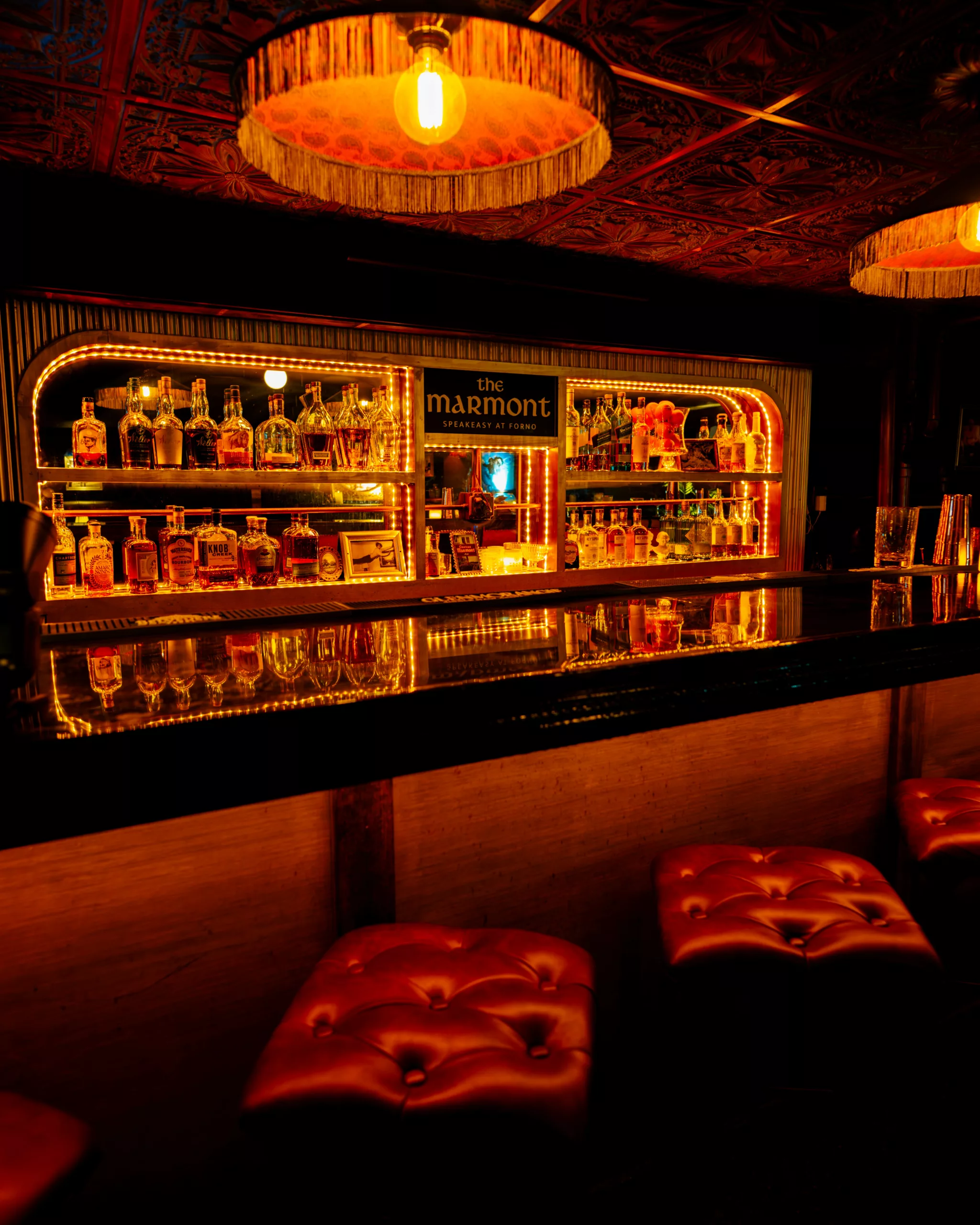 Speakeasy bar with lit shelves, a fringed lamp and tufted stools at Forno Kitchen + Bar