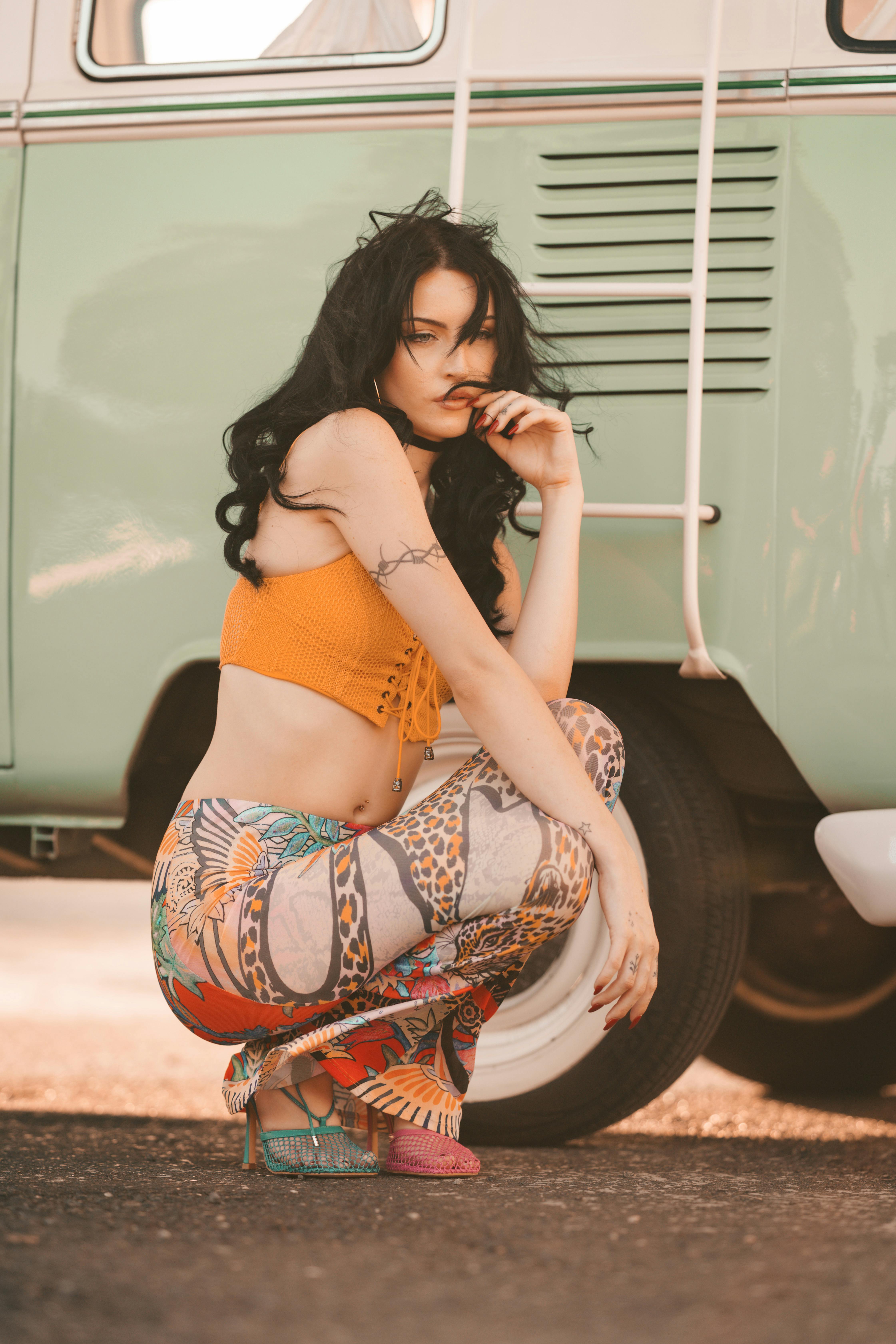 Stylish woman with long black hair posing in front of a vintage green van, wearing a bright orange crop top, patterned pants, and colorful heels.