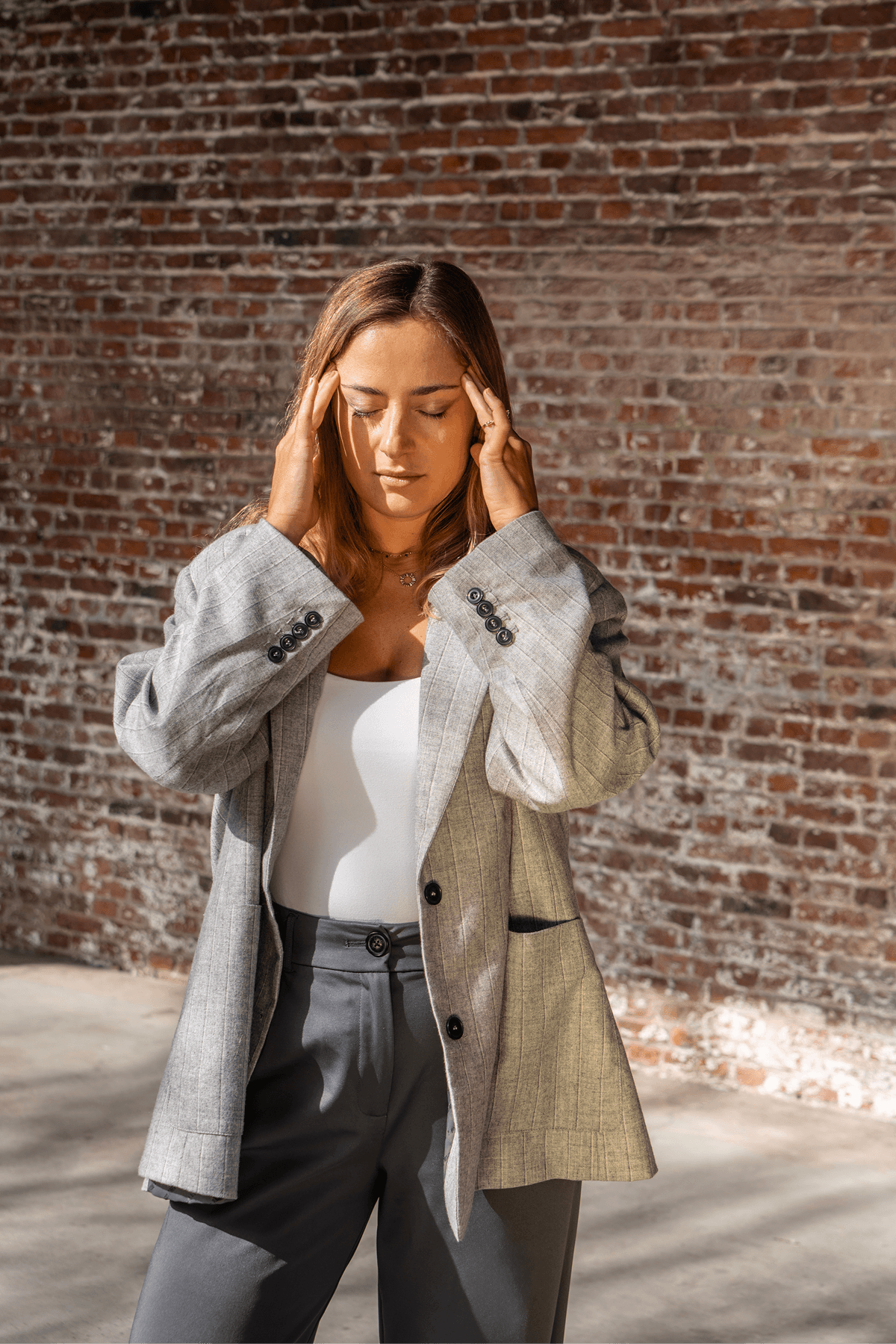 A person wearing a light gray blazer and dark pants stands against a textured brick wall, with eyes closed and hands gently pressing their temples, symbolizing stress and the need for burnout prevention.