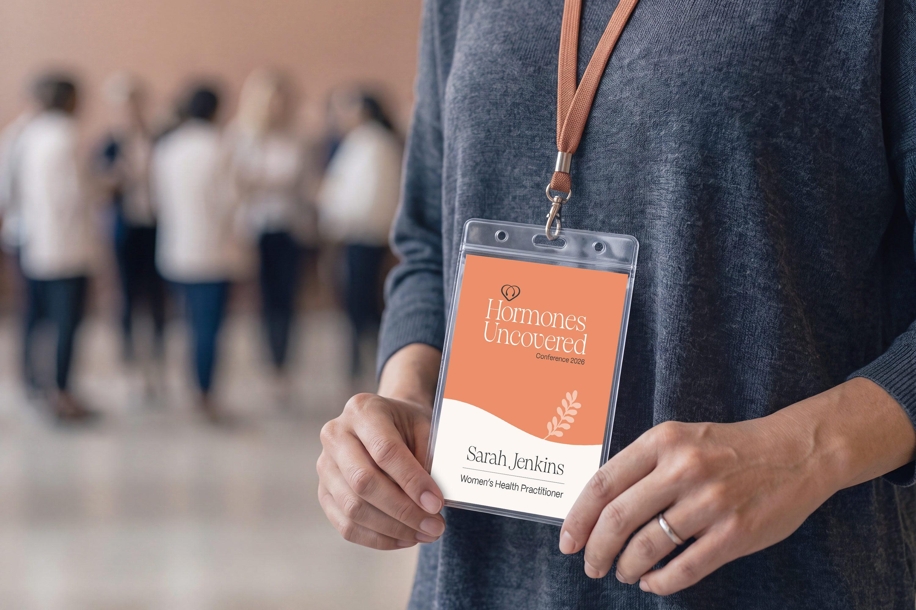 A woman holds a name badge, with a blurred group of people chatting in the background at an event.