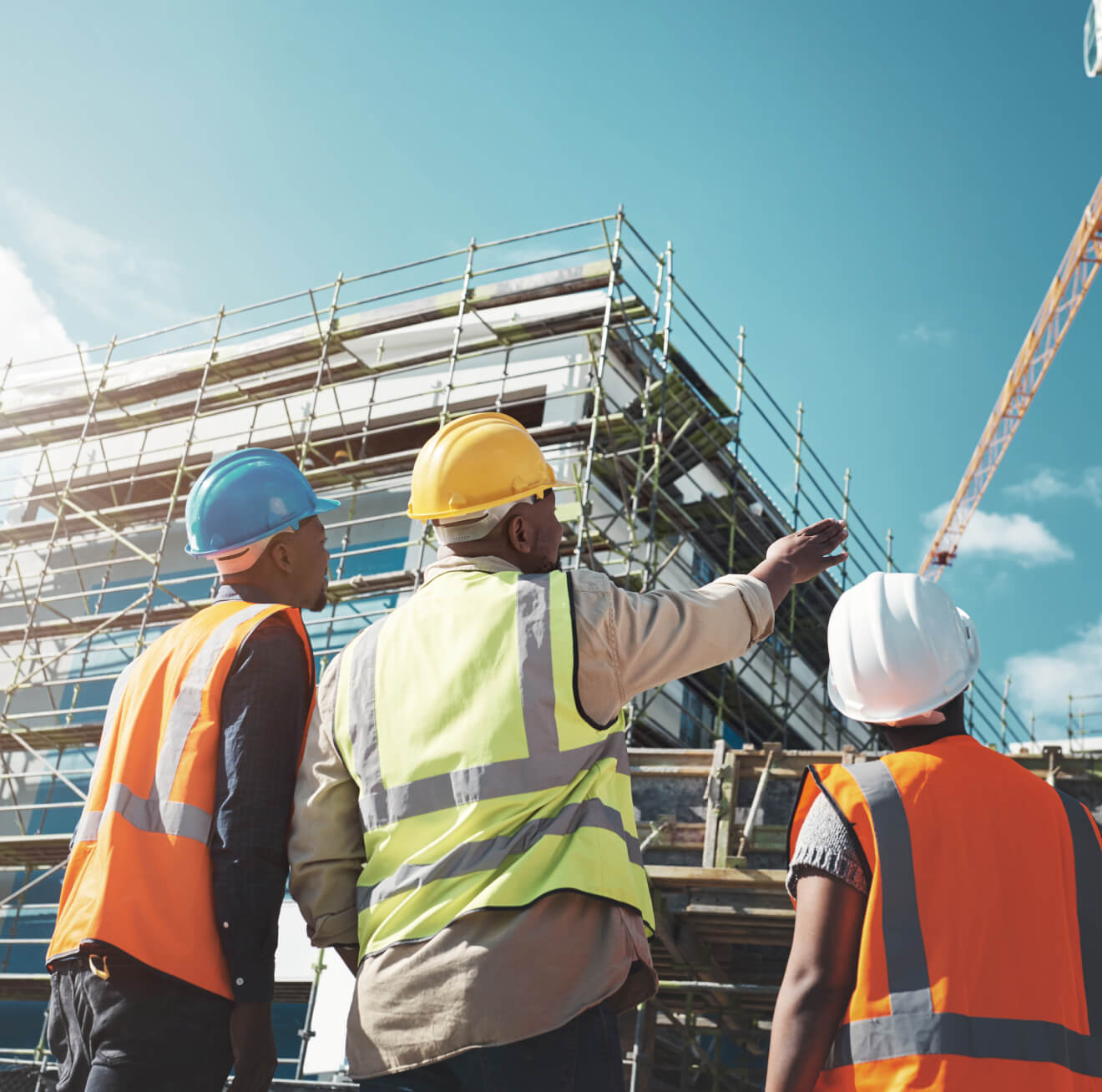 Three construction workers in safety gear observe a building under construction, pointing and discussing.