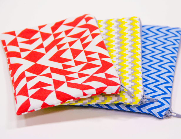 Three zippered pouches are stacked on a white surface. The pouch on the left is red with white triangles. The middle pouch is white with yellow zig-zag lines. The pouch on the right is blue with blue zig-zag lines.
