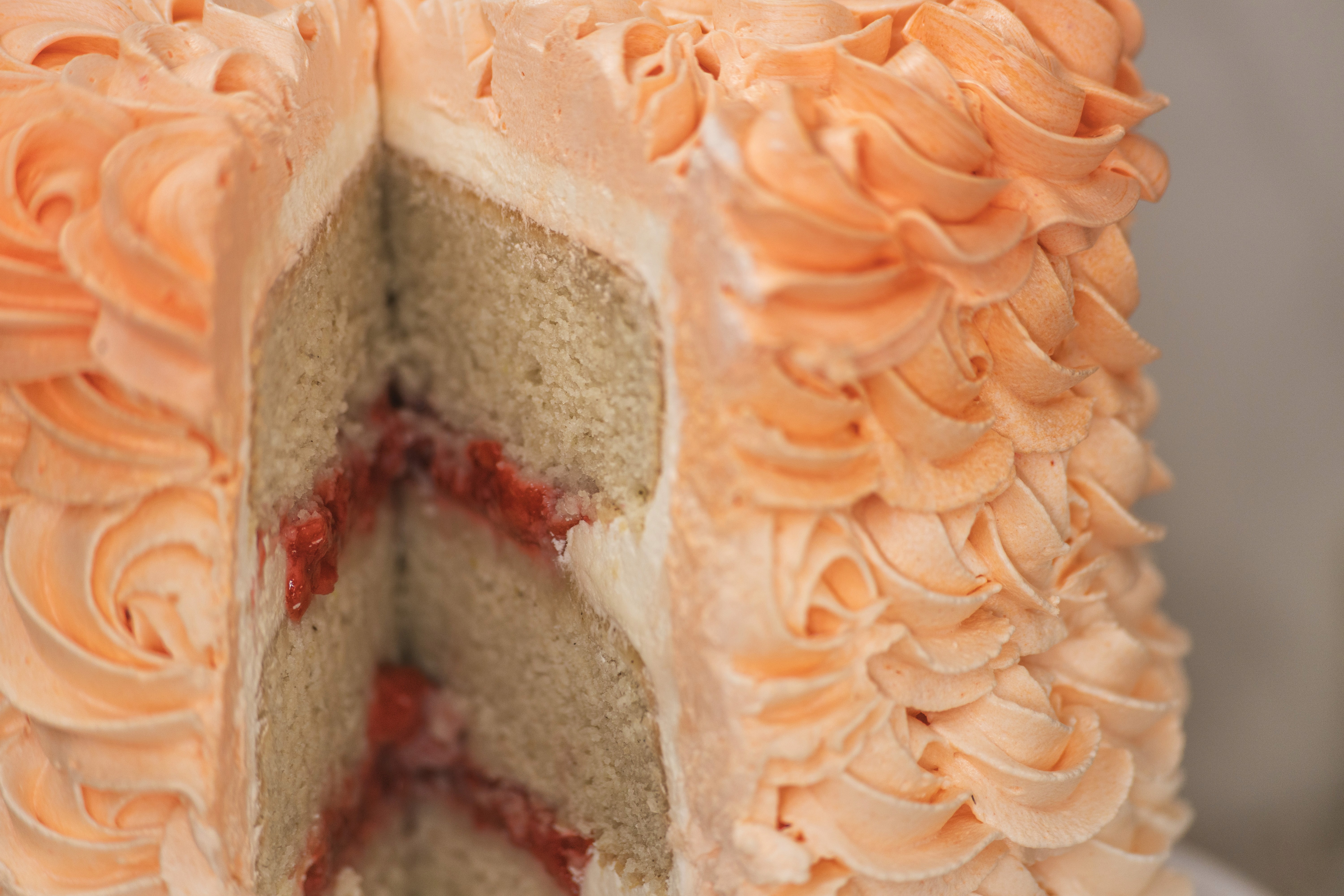Close-up shot of a tall layer cake slice featuring fluffy vanilla sponge layers, red jam filling, and pale peach buttercream frosting.