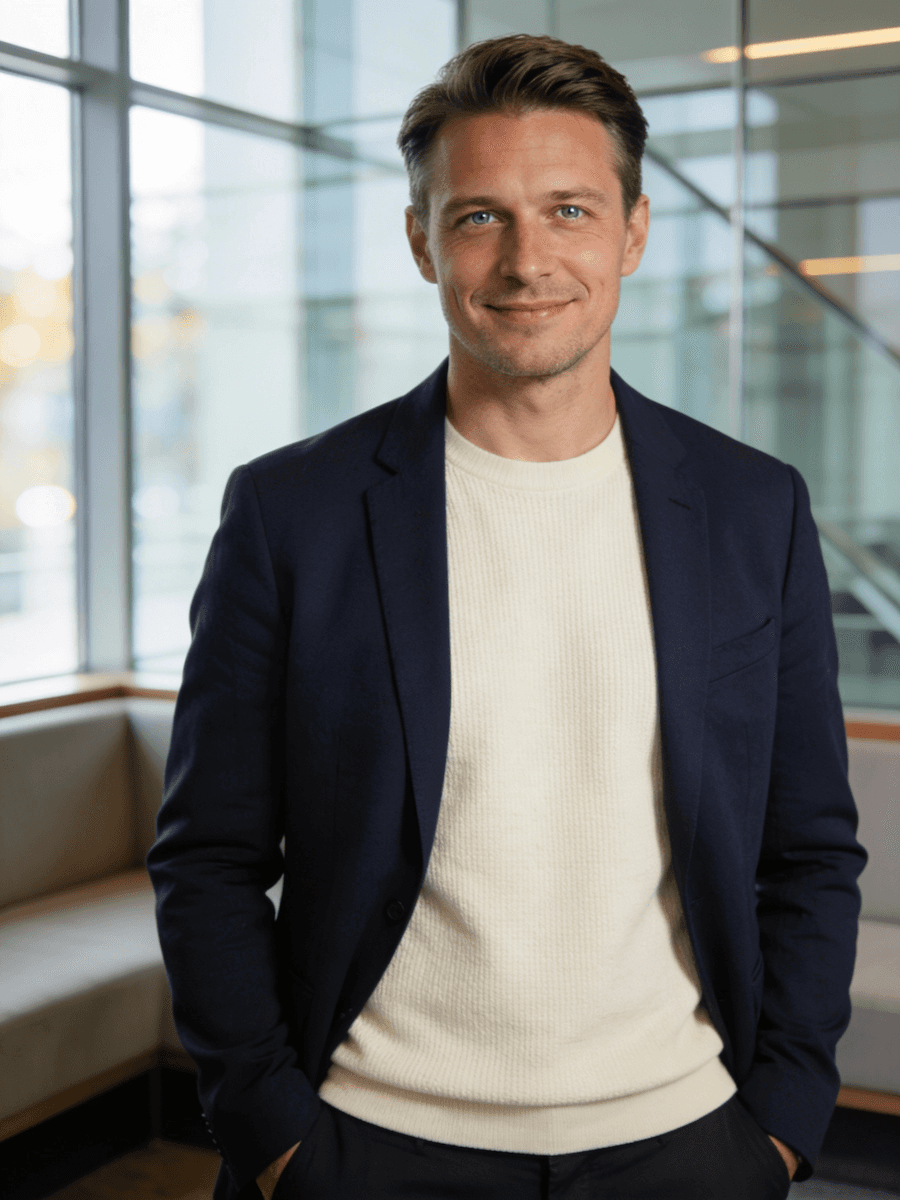 A smiling man in a dark jacket and light shirt stands in a bright, modern indoor setting.