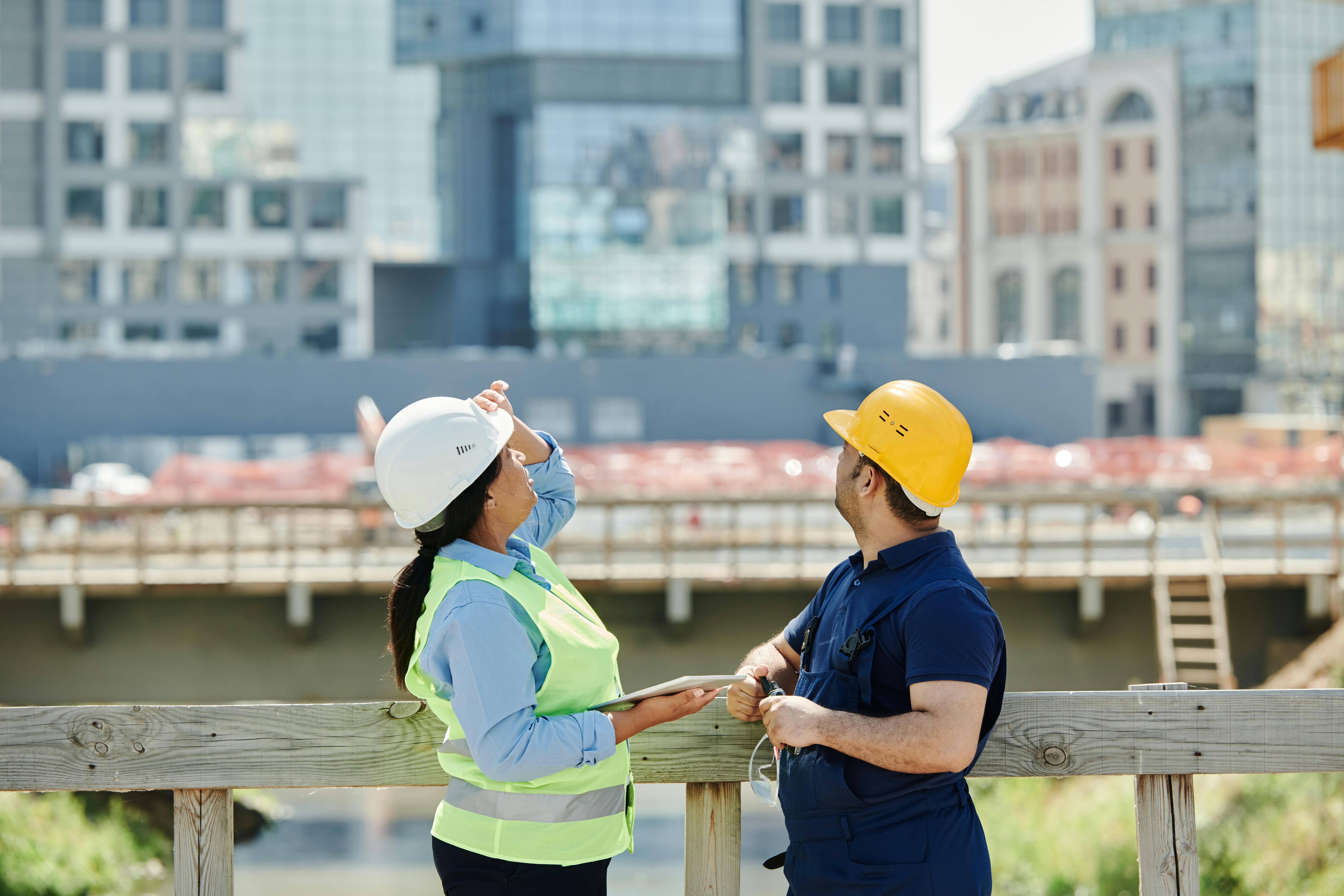 Two construction workers wearing safety vests and hard hats discuss work at a building site. A cityscape behind them underlines progress.