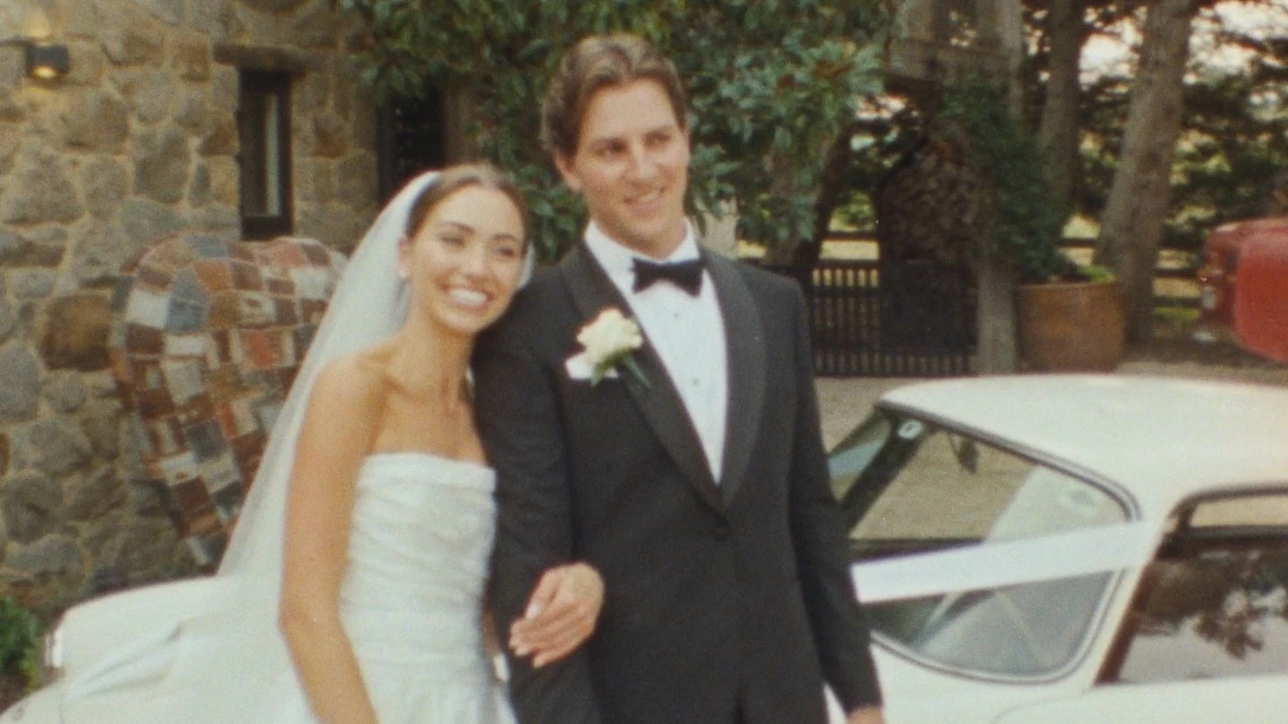 A bride in a strapless white wedding gown and veil stands smiling with a groom in a black tuxedo and bow tie, in front of a classic white car and stone building, surrounded by greenery.