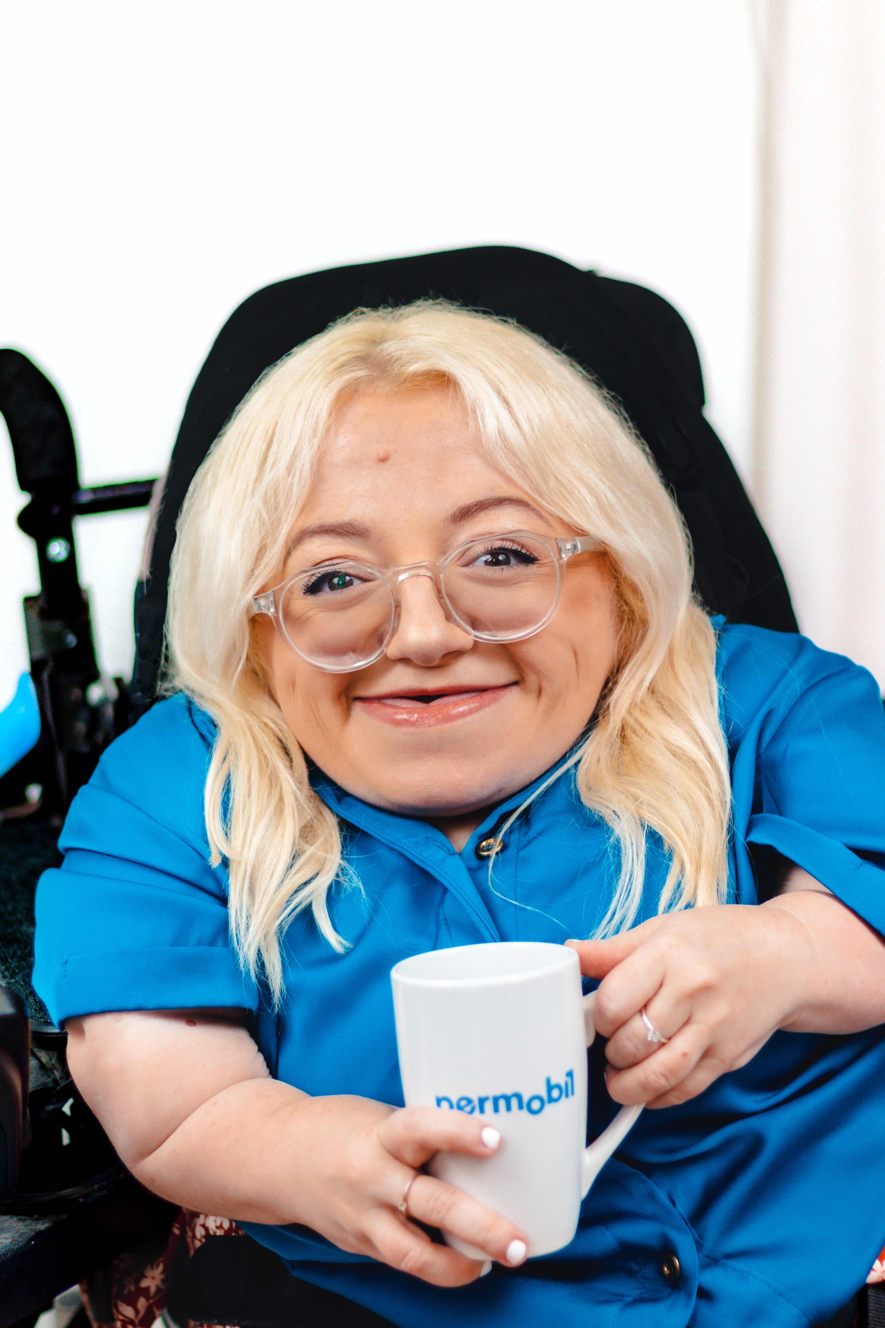 Gem is wearing a bright blue shirt and holding a Permobil mug. She has a big smile on her face.